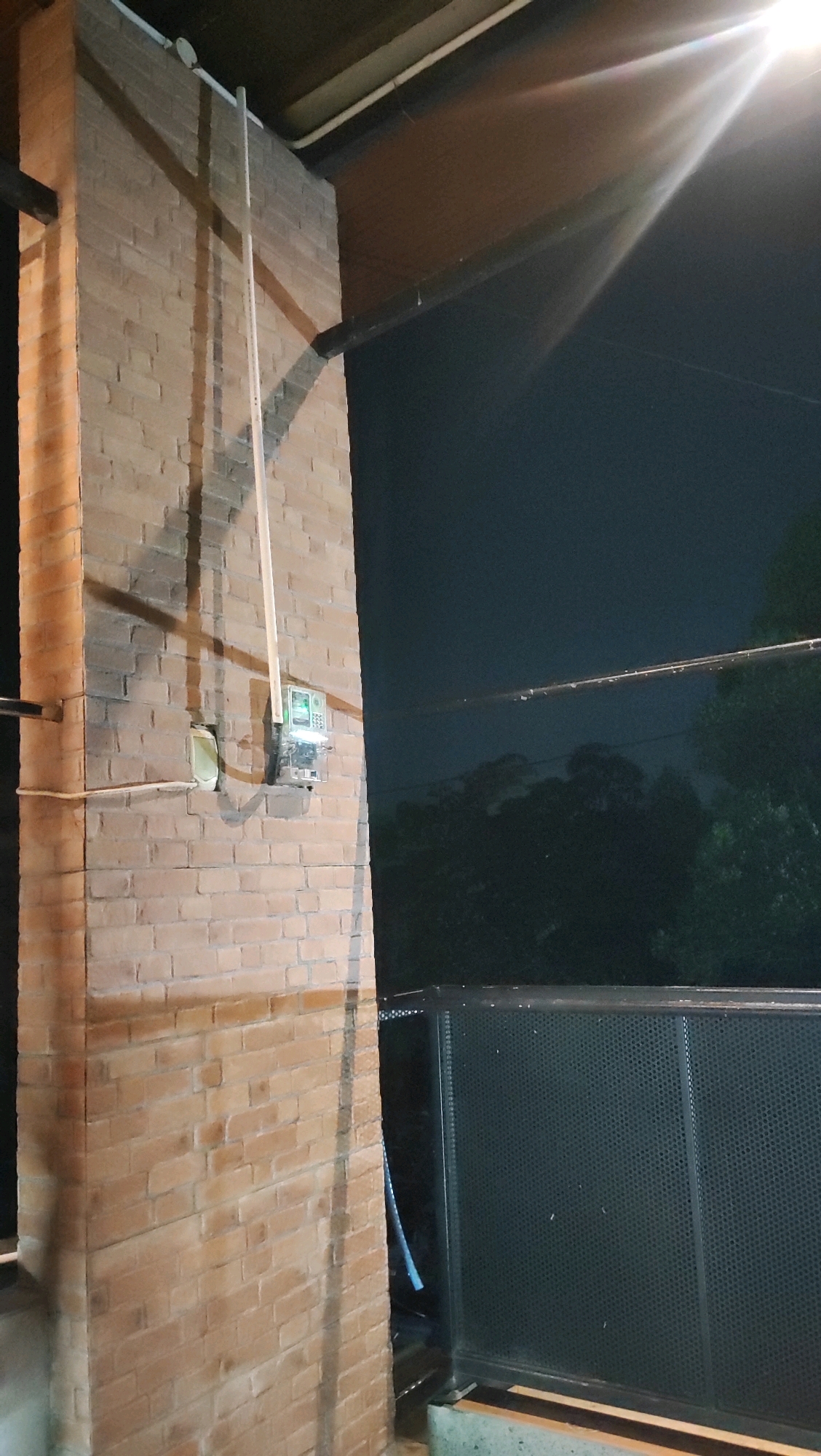 A nighttime outdoor medium shot shows a brick pillar with an electricity meter attached to it. The pillar is made of light brown bricks. A white conduit runs vertically up the pillar, terminating at the meter. The meter itself is silver with a green digital display. To the right of the pillar is a dark, perforated metal barrier. In the background, dark trees are visible against a black sky. A bright light source, possibly a streetlamp, casts a strong glare across the top right of the image. The scene appears to be in a residential or commercial area in Karangwangkal, Indonesia.
