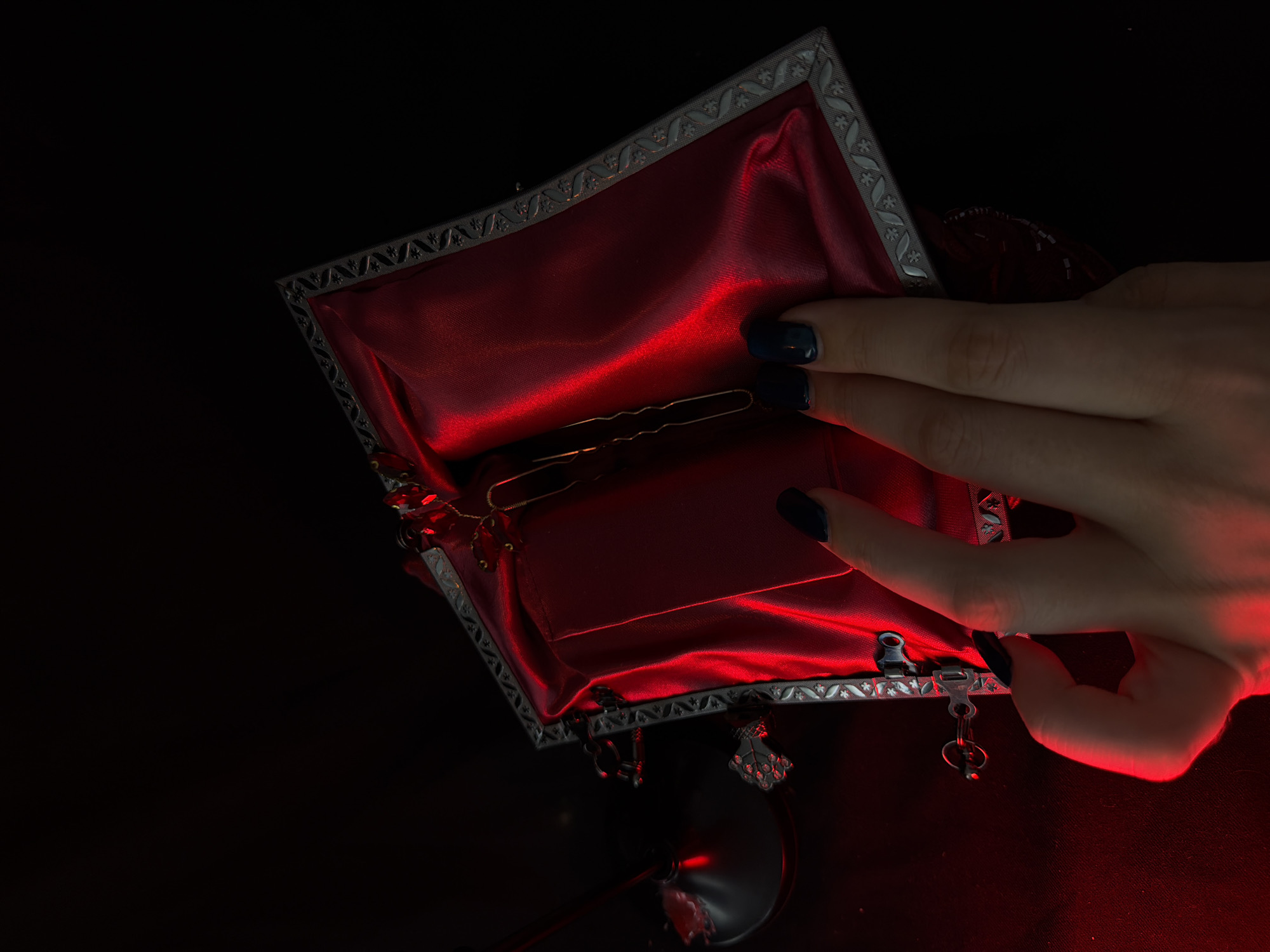 A close-up shot focuses on a hand with dark nail polish reaching into an ornate, open jewelry box lined with red satin. The box has a decorative silver trim and appears to be partially open, revealing a thin, gold-colored necklace or chain inside. The lighting is dramatic, with a strong red glow casting shadows and highlighting the textures of the satin and the hand. In the background, a dark, out-of-focus element, possibly a glass or stand, is also illuminated by the red light. The overall impression is one of luxury and mystery.