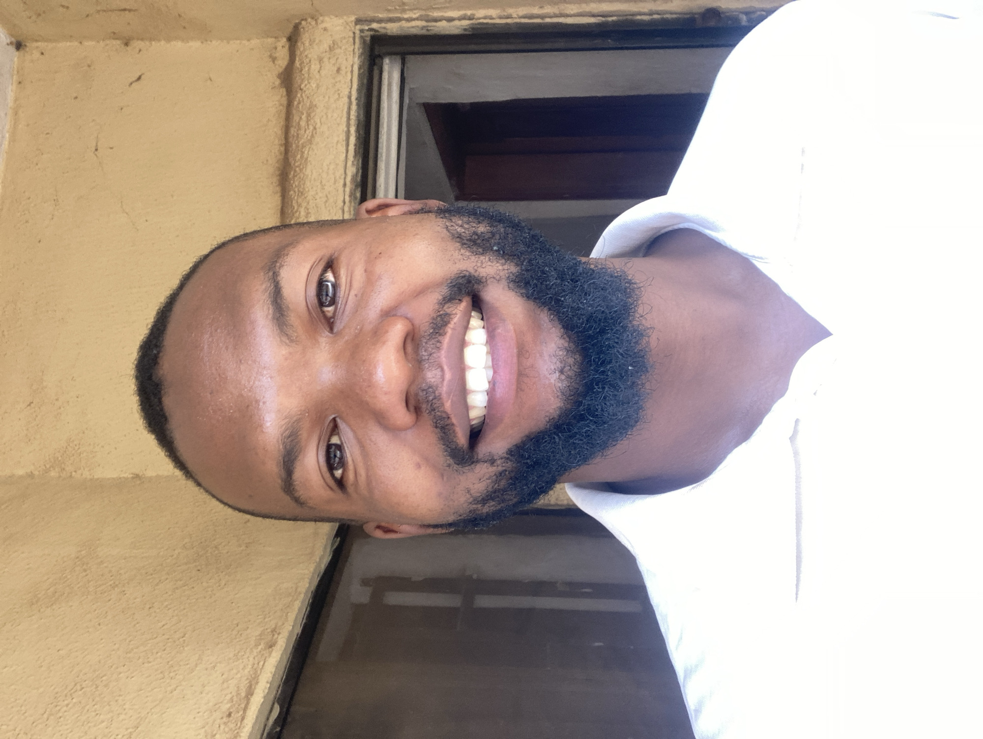 A close-up portrait shows a smiling man with dark skin, short dark hair, and a full black beard. He is wearing a white shirt. The background includes a textured, tan-colored wall and a dark window frame with a glimpse of a dark interior. This image was likely taken in Jos, Nigeria.