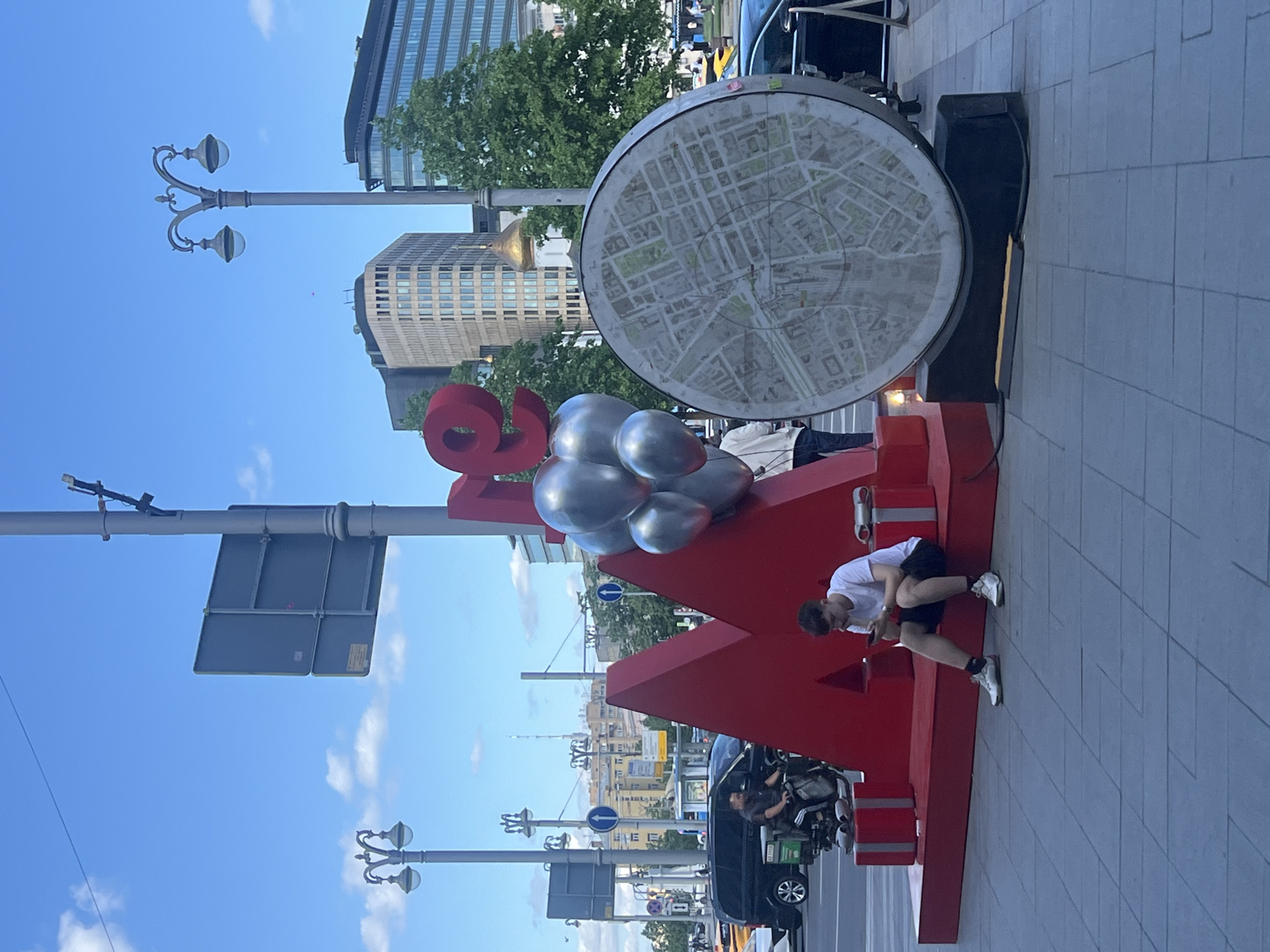 A pedestrian is sitting on a large, red "Я люблю Москву" (I love Moscow) sculpture located in Moscow, Russia. The sculpture has several silver, balloon-like shapes placed on top. Behind the sculpture is a circular street map, as well as several buildings and trees. There are a variety of vehicles in the road. The sky is blue and the weather appears sunny.