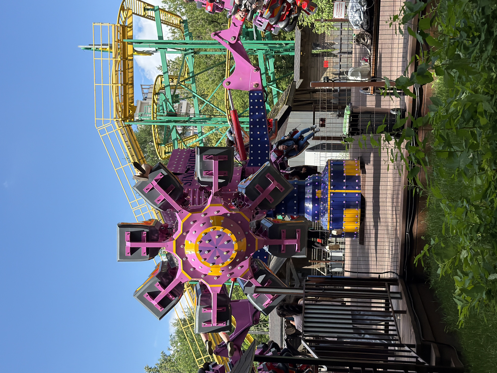 The image shows an amusement park ride with people on it. The ride is predominantly purple and yellow with some blue accents. It appears to be a spinning or rotating ride with individual carriages. In the background, a yellow and green roller coaster track is visible against a bright blue sky. Trees and foliage surround the area, suggesting an outdoor setting. The lighting suggests it is daytime, likely a sunny day given the clear sky. There are also safety fences and wooden platforms typical of an amusement park.
One of the carriages on the main ride has text visible on its side, which appears to be in Cyrillic script. Part of the text reads "ОСТОРОЖНО ЗАПРЕЩЕНО" which translates to "CAUTION FORBIDDEN." This indicates a warning or restriction related to the ride or its operation. The people on the ride appear to be enjoying themselves, with some smiling and looking excited. The overall mood seems to be one of fun and exhilaration, characteristic of an amusement park visit. The location context of Perm, Russia, places this scene within that specific geographical area.