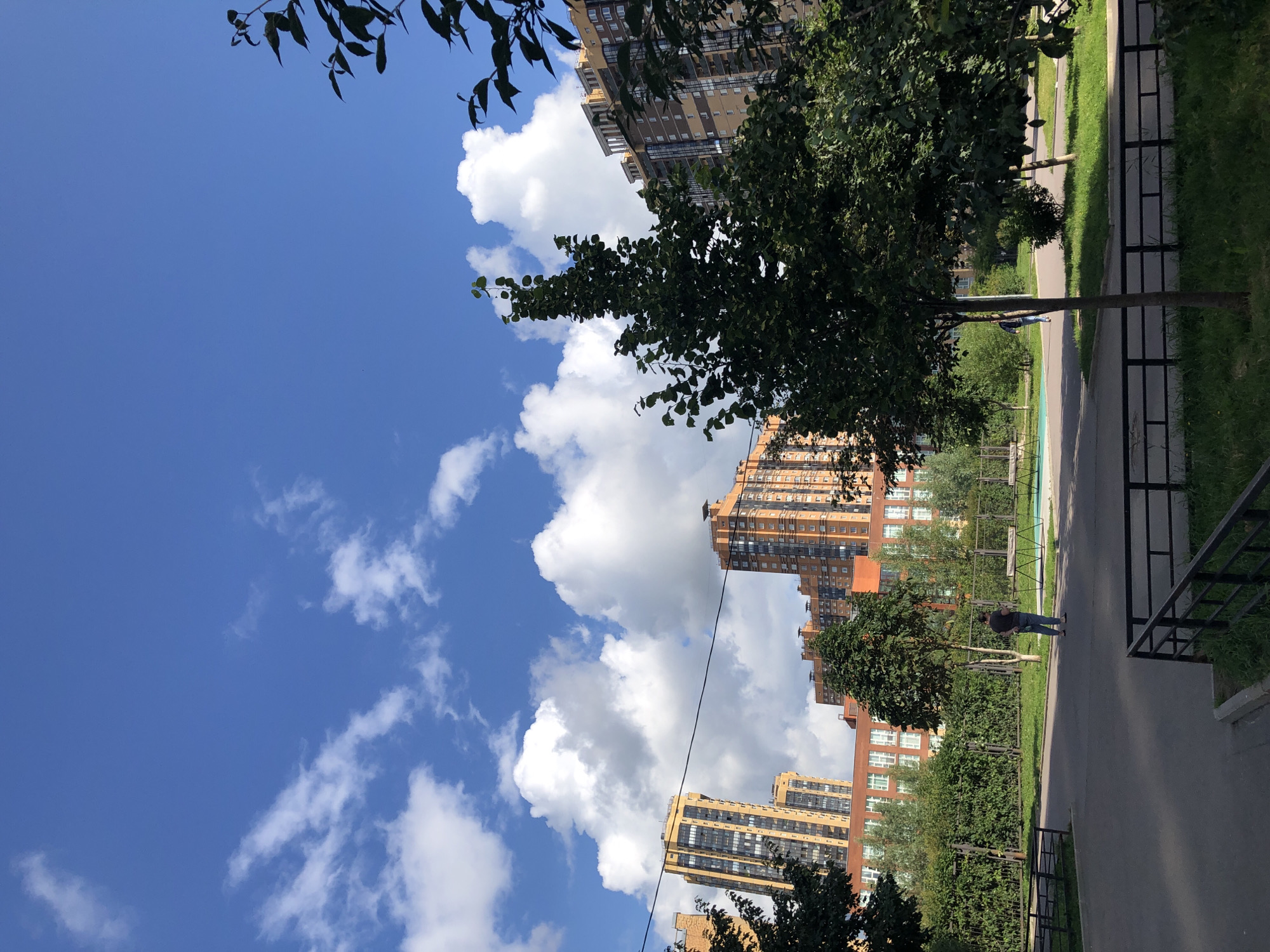 The image depicts an outdoor urban scene with several high-rise buildings in the background, characterized by their orange and yellow facades. The sky is a vibrant blue with scattered white cumulus clouds. In the foreground, there is a paved pathway and a grassy area with some trees. A few people are visible; one person stands in the middle ground facing away from the camera, and another person walks in the distance. A black metal fence runs along the edge of the pathway. The scene appears to be a residential area, possibly a courtyard or park, with playground equipment visible in the distance. The lighting suggests it is daytime, likely sunny.
