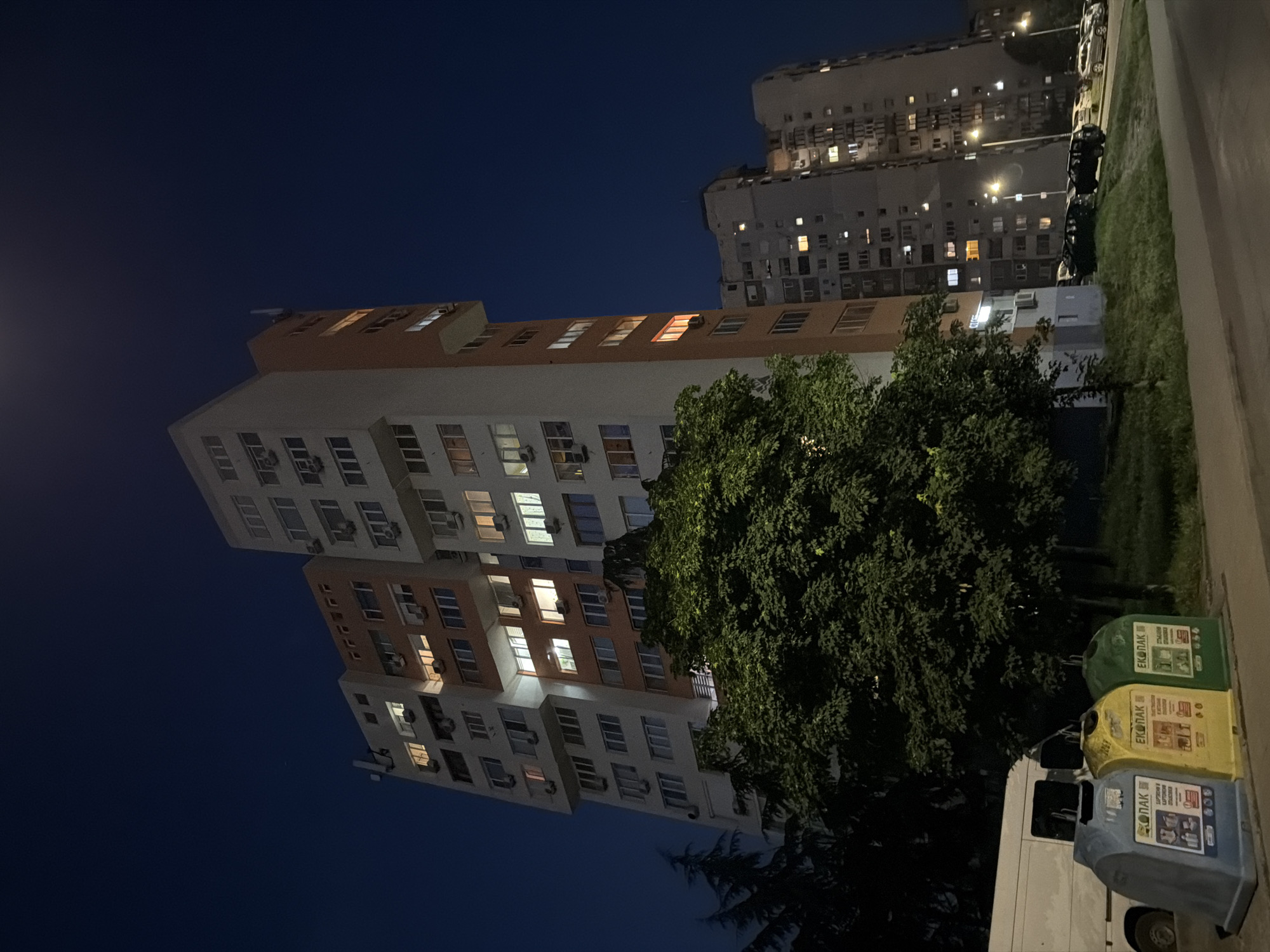 The image depicts a nighttime urban scene in Burgas, Bulgaria, specifically at the residential complex located at Slivnitsa Street 17A, 8000 Burgas Center. The dominant feature is a tall, multi-story residential building positioned centrally, characterized by a segmented facade with sections of white and orange/terracotta panels. Numerous windows are visible across all floors; some are internally illuminated, casting light, while others remain dark. Multiple external air conditioning units are mounted beneath various windows.
In the foreground, on the left side of a paved road, are three large, distinct recycling bins identified by the "ЕКОПАК" (ECOPACK) branding: a blue bin designated for plastic and metal, a yellow bin for paper, and a green bin for glass. Partially obscured behind these bins is the rear section of a white commercial van. A large, leafy deciduous tree with dense foliage occupies the central foreground, partially concealing the lower levels of the main building.
To the right, a row of parked vehicles, including several SUVs and sedans, lines the side of the paved road. The street and vehicles are illuminated by multiple modern streetlights. In the background, additional multi-story residential buildings with numerous illuminated windows are visible, contributing to the dense urban environment. The sky is dark blue and clear. No people are visible within the frame.