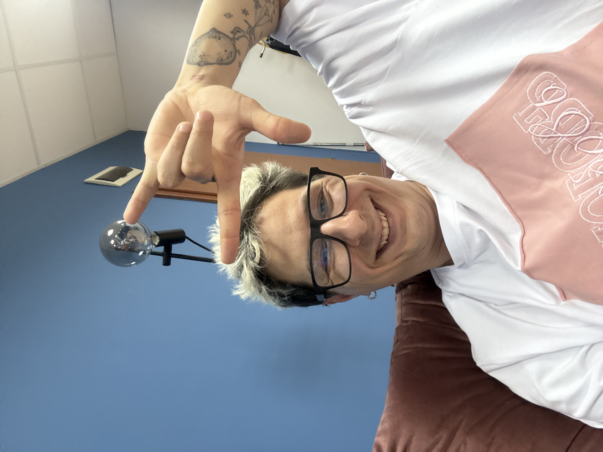 A male subject, with short, spiky hair bleached blonde over dark roots, smiles broadly directly at the camera. He wears black-framed glasses and a small silver hoop earring in his left ear. His right arm is raised, with his hand forming a rectangular framing gesture; his thumb and index finger are extended to create an 'L' shape, while his remaining fingers are curled. Visible on his right forearm are multiple tattoos, including a grey, circular, possibly planetary or lunar design, and several smaller, linear star or floral motifs. A small red mark or scar is present on the inner aspect of his right wrist. He wears a white short-sleeved t-shirt featuring a distinct pink fabric panel on the lower right, embroidered with white text, partially legible as "SOFIA" in block letters and additional cursive script.
The subject is seated on what appears to be a reddish-brown upholstered chair or sofa, partially visible on the left. The background consists primarily of a solid blue wall. On the blue wall, behind the subject's right shoulder, a white-framed, diamond-shaped object is mounted. Above the blue wall, a white suspended ceiling with a visible grid pattern is present. A modern light fixture with a spherical clear bulb and a black stand extends into the frame from the left, positioned behind the subject's head. The indoor environment suggests an office, studio, or common room setting. The word "SOFIA" is present on the subject's apparel.