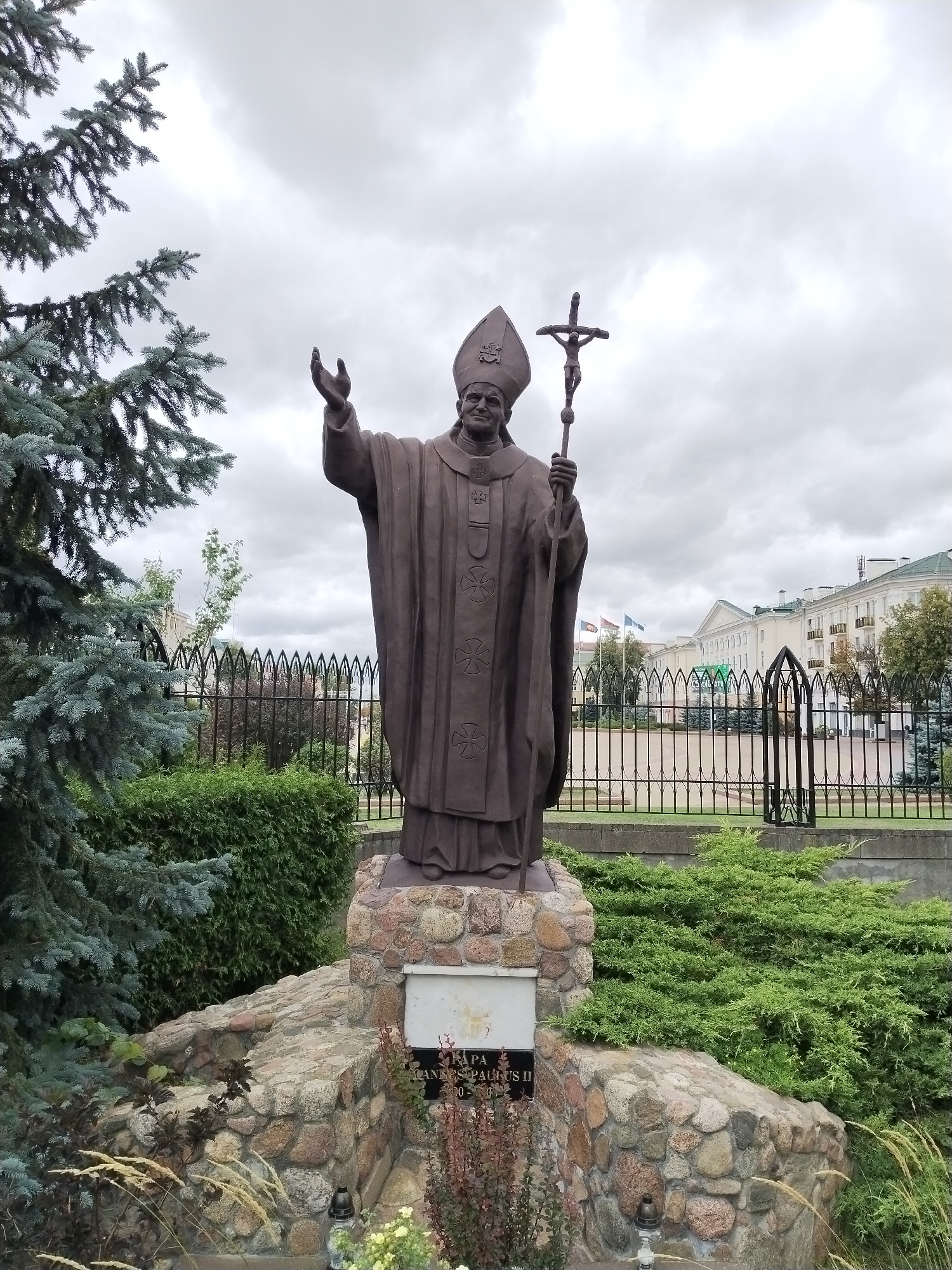 A full-body bronze statue of Pope John Paul II stands on a stone pedestal in a public space. The Pope is depicted wearing his papal vestments and tiara, holding a crucifix in his right hand and with his left hand raised in a blessing gesture. He is looking downwards with a gentle expression.
The statue is situated in front of a black metal fence, behind which a row of green bushes and trees are visible. In the background, a multi-story building with a light-colored facade, green roof, and balconies can be seen. Several flags are also visible in front of the building.
The foreground includes a stone base for the statue, adorned with smaller rocks and low-lying green shrubs. There are also some ornamental grasses and a memorial candle placed on the stone base. To the left of the statue, a large blue-green conifer tree is prominently featured. The sky above is overcast with grey clouds.
The inscription on the memorial plaque at the base of the statue reads "PAPIEŻ JAN PAWEŁ II" and the dates "1920-2005." This suggests the statue is a memorial to Pope John Paul II.
The location is the area in front of the Roman Catholic Church of the Exaltation of the Holy Cross in Brest, Belarus, where a monument to Pope John Paul II was erected.