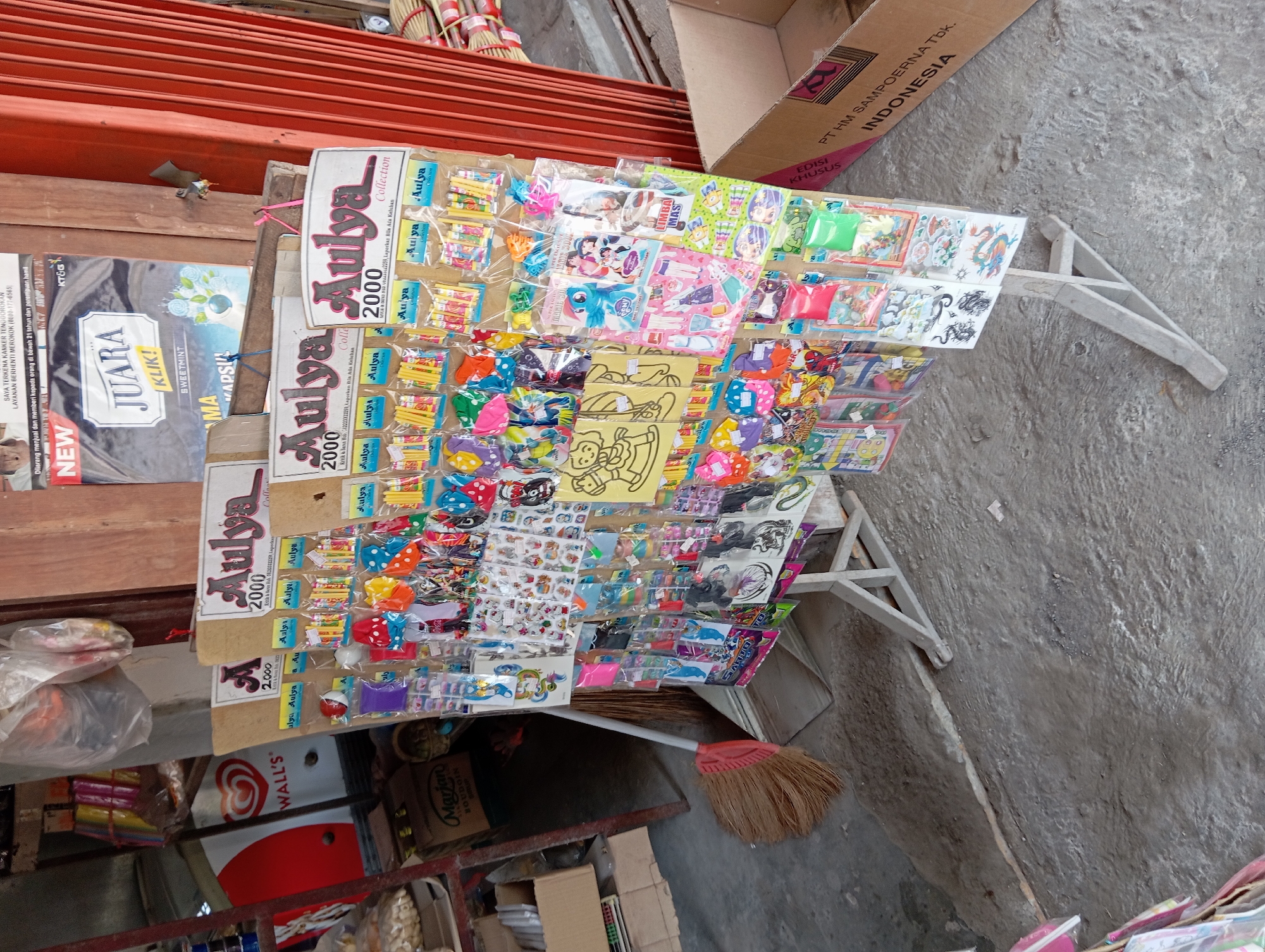 The image depicts an outdoor stall, likely in the city of Srumbung, Indonesia, displaying a variety of small toys and novelty items. The stall is constructed with a light-colored wooden frame and is filled with an assortment of individually packaged items, predominantly stickers, miniature games, and colorful accessories. These items are neatly arranged on cardboard backing and hung from the display.
Prominently featured are multiple signs with the brand name "Aulya," often accompanied by a price of "2000." The items themselves are diverse, including what appear to be temporary tattoos, small puzzles or games, and various decorative stickers featuring cartoon characters, animals, and traditional motifs like dragons. The packaging is primarily clear plastic, allowing the colorful contents to be visible.
In the background, a red corrugated metal door or wall is visible, suggesting the stall is positioned against a building. To the left of the stall, there's a glimpse of a refrigerator with a "WALL'S" logo, indicating it might be a food or beverage vendor nearby. Further to the left, a broom with a reddish-orange handle and straw bristles rests against a stack of cardboard boxes. One box is labeled "Mariani BOURGOIN." A poster with "NEW JUARA KLIK!" is also visible on the background wall. The ground is a rough, gray concrete surface, suggesting a street or walkway. There are no people visible in the image.