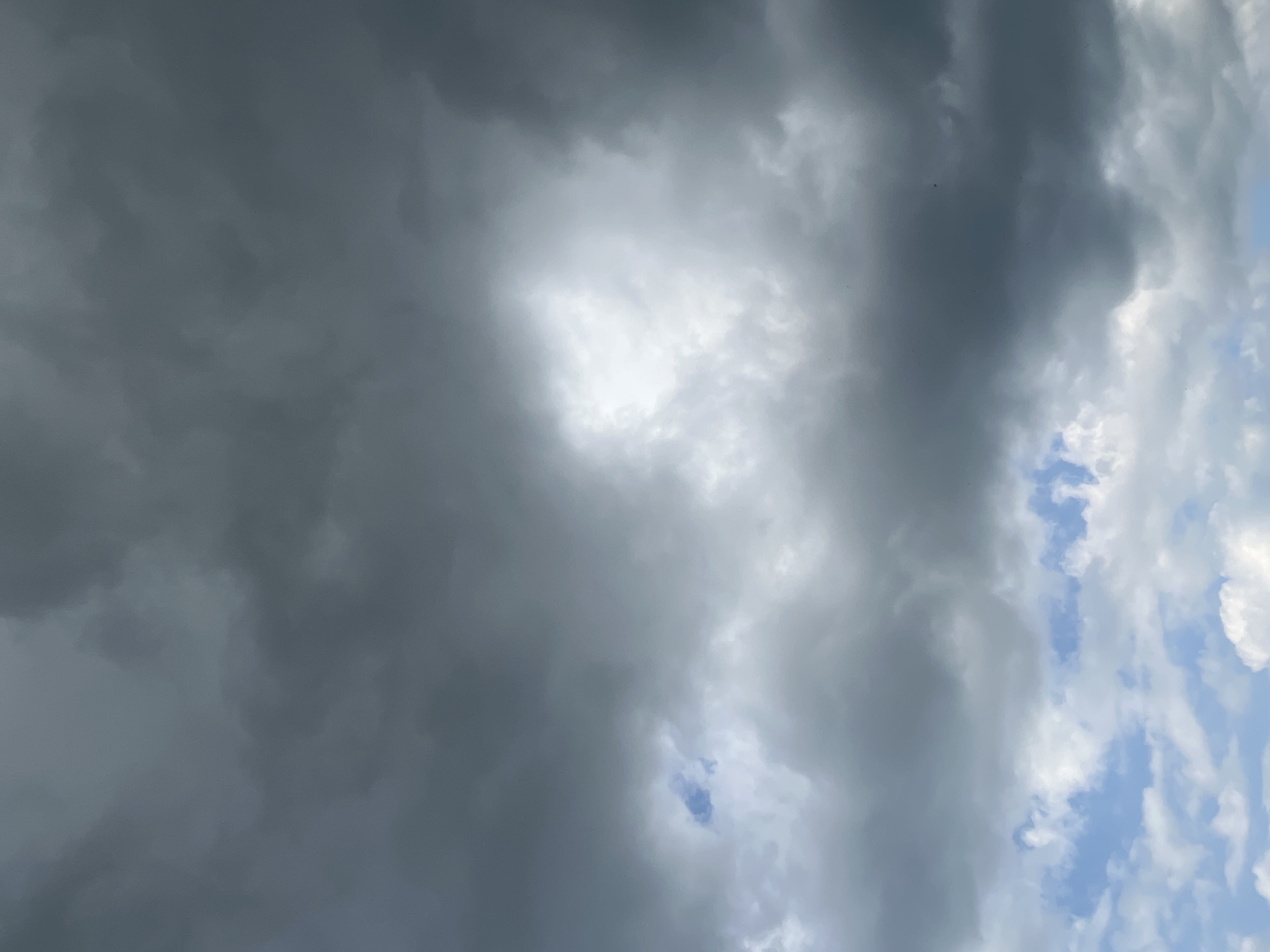 This is a photograph of a cloudy sky with patches of blue visible. The clouds are dense and dark in some areas, suggesting an approaching storm or overcast weather. In the lower part of the image, lighter clouds and patches of blue sky are more prominent. There are no people, objects, or identifiable landmarks in the image, making it impossible to determine a specific location within Il'ino, Russia or any other city. The focus is solely on the atmospheric conditions.
