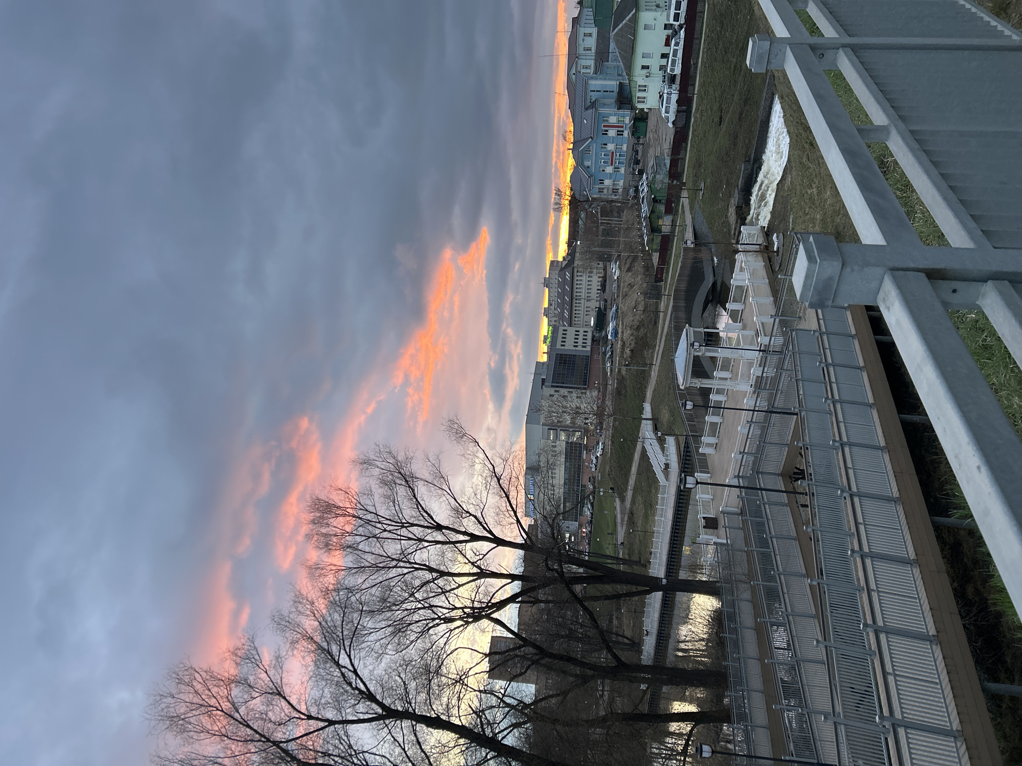 The image shows a cityscape during twilight or early morning, possibly in Klin, Russia. The composition is dominated by a dramatic sky with a mix of dark grey and pink-tinged clouds. Bare trees are silhouetted in the foreground.
In the middle ground, a mix of buildings is visible. These buildings include modern high-rises and older, possibly residential, structures. A paved area with white structures, possibly part of a park or public space, runs through the scene. A narrow body of water, likely a river or canal, reflects the sky and buildings.
A metal railing and support structures run along the right side of the frame, suggesting the image was taken from an elevated vantage point, such as a bridge or building. The vegetation on the ground indicates it might be early spring, with patches of snow still visible.