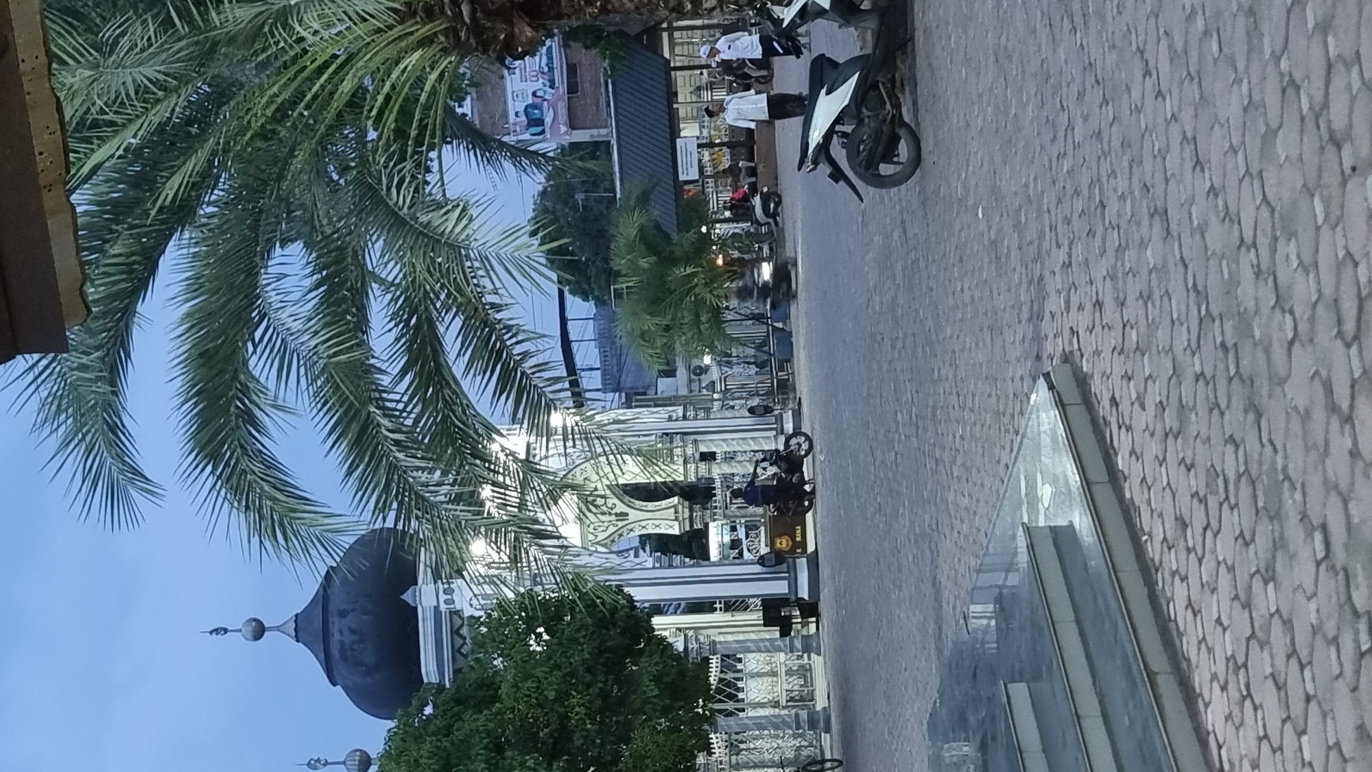 The image depicts the exterior of a mosque, likely in Keudekruenggeukueh, Indonesia, with a paved courtyard in the foreground. A large palm tree is prominently featured, with its fronds partially obscuring the sky. The mosque has a dark dome and intricate white architectural details, including arches and columns. Several people are visible in the courtyard, some standing and others on motorcycles. One individual is near a motorbike in the foreground, while others are further back, near the mosque entrance or seated under a covered area. The scene appears to be set during twilight or early evening, with artificial lights illuminating the mosque. Several motorbikes are parked or in motion within the courtyard. The ground is paved with interlocking hexagonal stones, and a set of tiled stairs is visible on the left side of the image. The overall atmosphere suggests a public space associated with a religious institution.