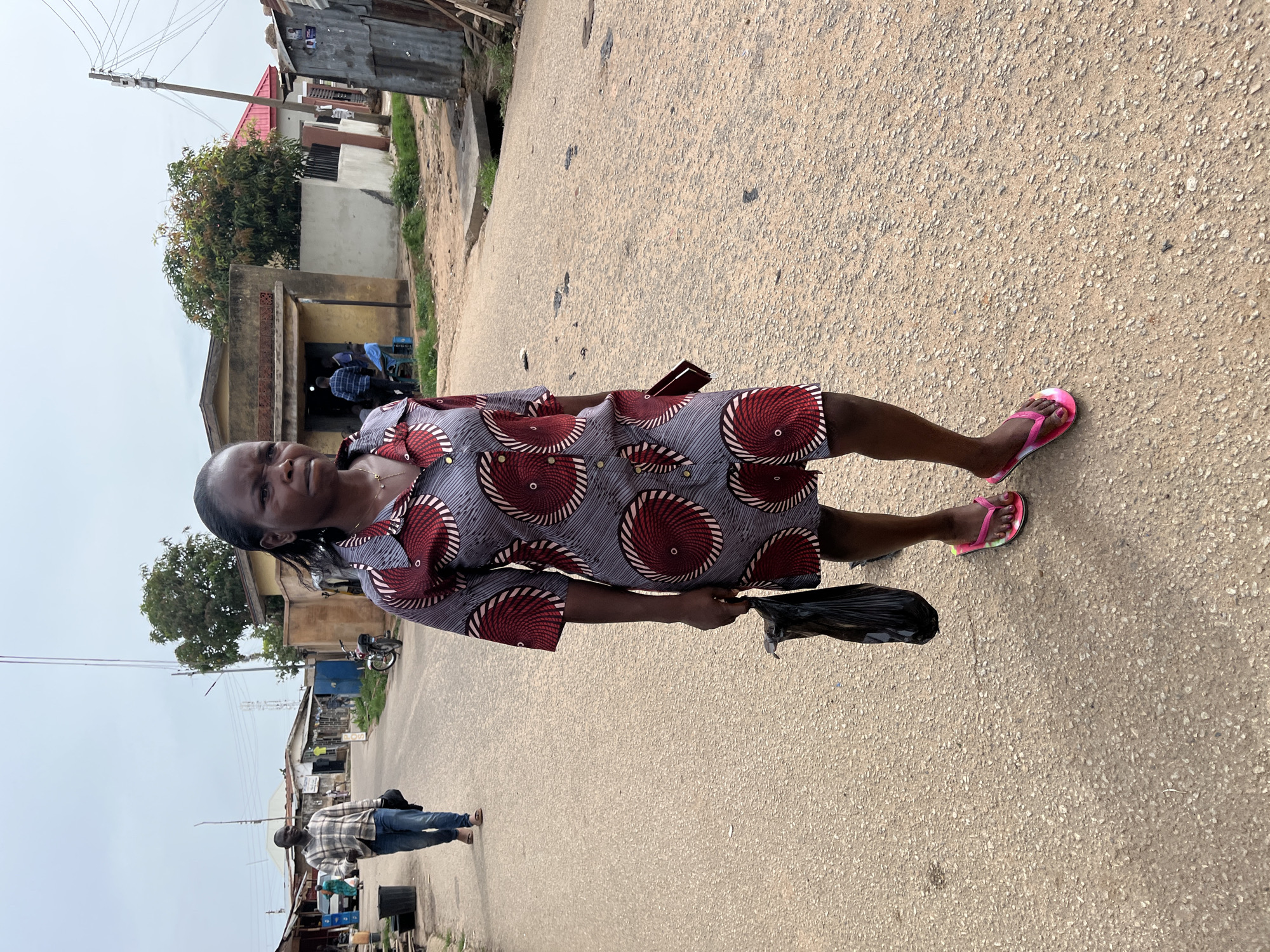 A woman wearing a patterned dress and pink flip-flops walks on a paved street in Jos, Nigeria. She holds a black plastic bag in her left hand. The environment includes buildings, some of which appear to be shops, a utility pole, and other people walking in the background.