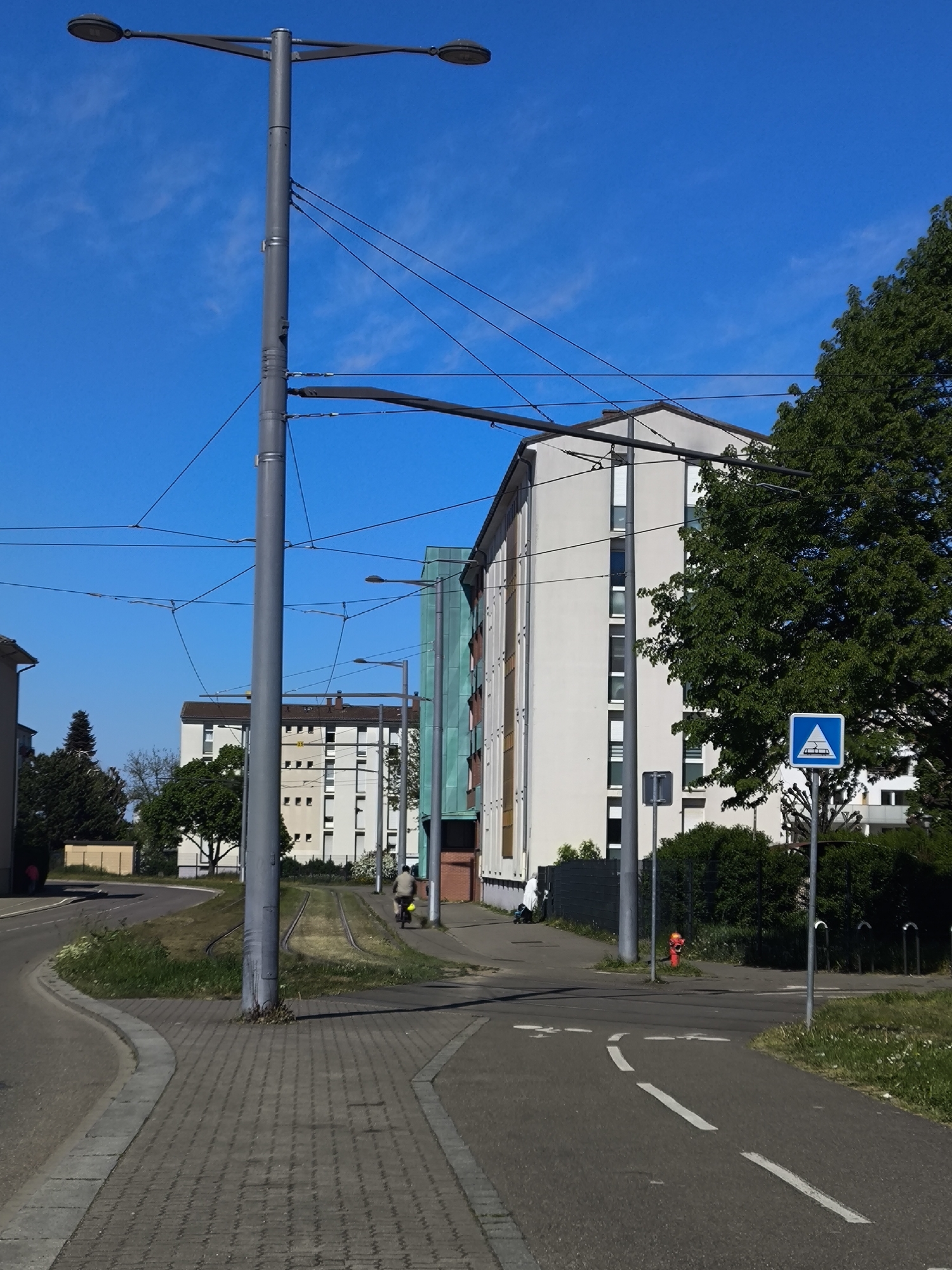 The image captures a street scene in Hoenheim, France, under a clear blue sky. In the foreground is a paved walkway and a bicycle lane marked with dashed white lines. Tram tracks run parallel to the walkway. A tall grey lamppost stands prominently, supporting overhead tram wires. Behind, several multi-story residential buildings are visible. To the right is a green tree, partially obscuring a blue and white sign with a tram icon. A red fire hydrant is visible near the crosswalk. A person on a bicycle and another person walking appear along the tram tracks. The surroundings consist of grass, hedges, and additional street furniture.