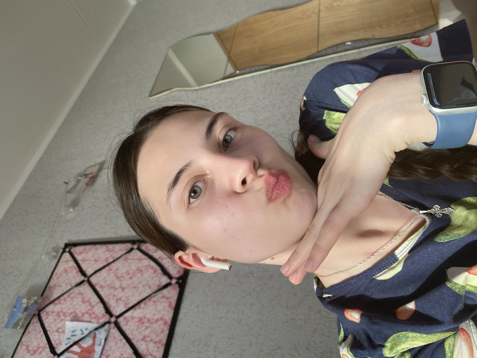 A woman poses indoors, facing the camera. She wears a navy-blue pajama top with a fruit pattern. A silver necklace with a cross pendant is visible. She has an Apple watch with a blue wristband on her left wrist. She has light skin, brown hair pulled back, and is wearing a white wireless earbud in her left ear. Her lips are pursed in a "kiss" expression. In the background, there's a wall with light-grey wallpaper, a multi-paneled mirror, and a patterned floor mat on the floor. There is not enough information to determine if the image was taken in Votkinsk, Russia.