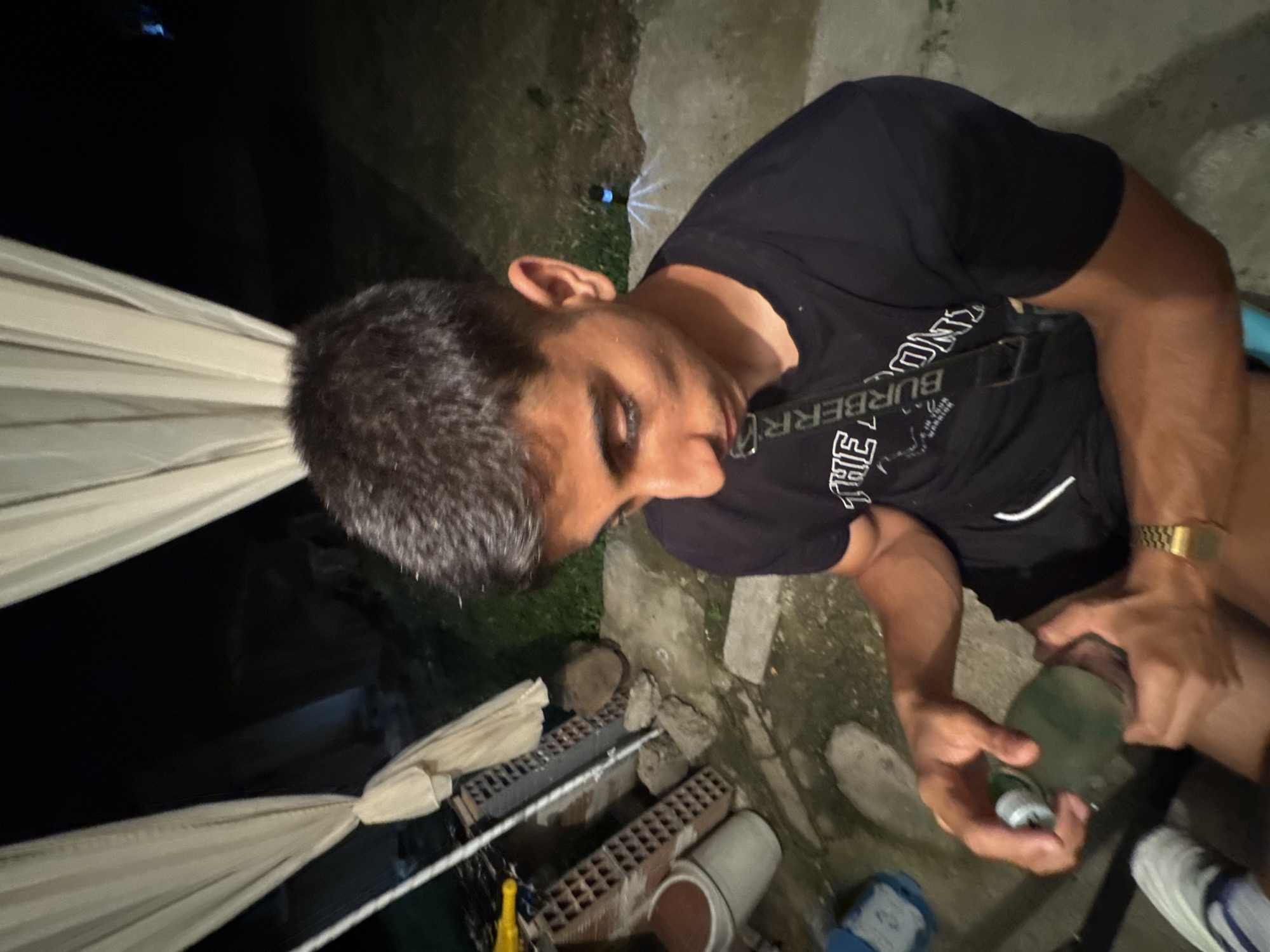 The image shows a young man with dark hair and a black t-shirt with white lettering and a strap across his chest. He is holding a green bottle in his hands. He is sitting on the ground outdoors at night. In the background, there are white curtains, some brick structures, and a small blue light from a solar-powered lamp. The ground appears to be made of concrete and dirt. The location is described as being in the city of Sredets, Bulgaria.