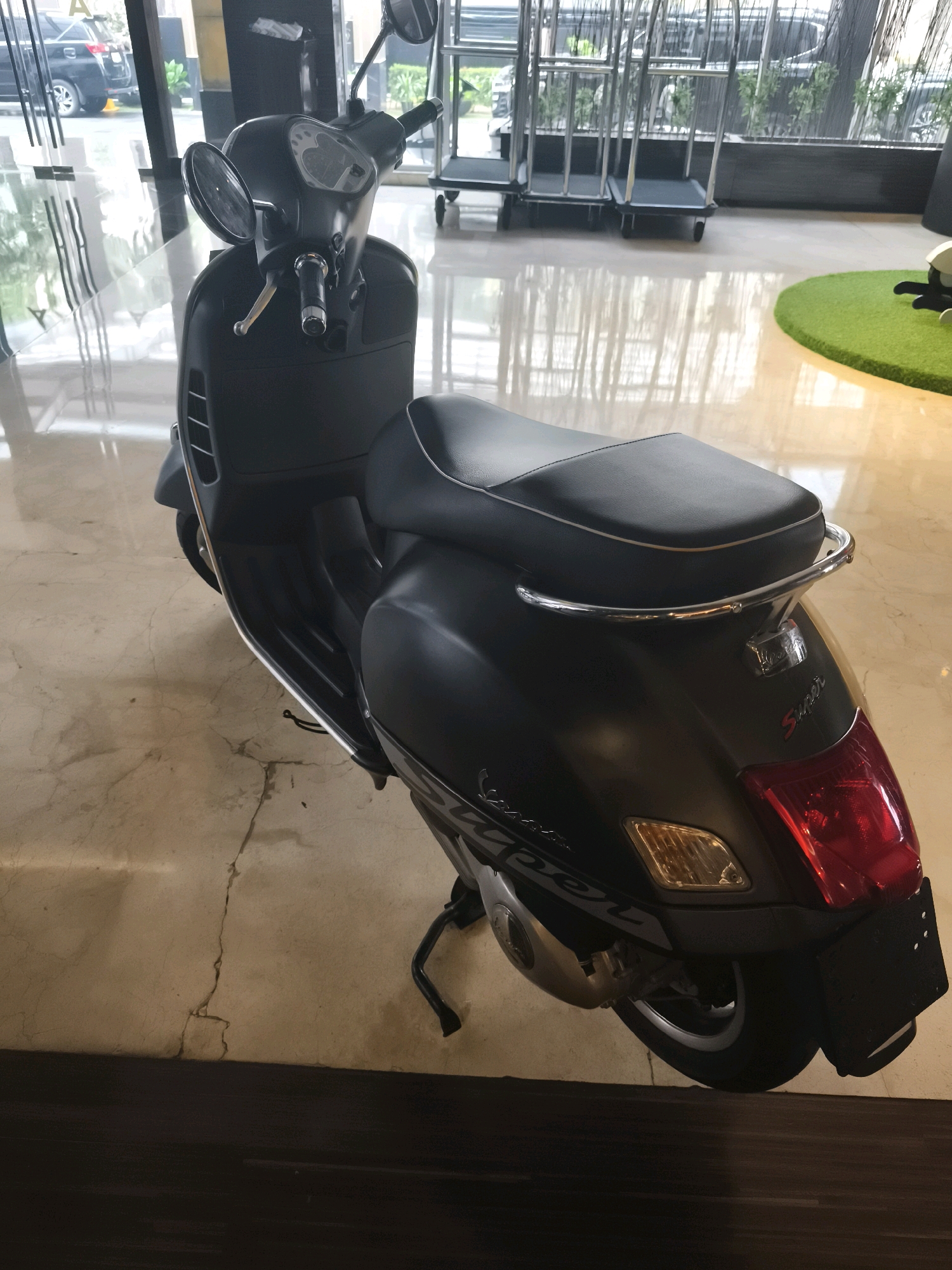 A black Vespa scooter is parked indoors on a polished marble floor, with a dark wooden border at the bottom of the frame. The scooter is angled to the right, showing its rear and side profile. It has a black leather seat with silver piping, a chrome grab handle, and a red taillight. The "Super" logo is visible on the rear bodywork. In the background, there are large windows showing an outdoor area with greenery and what appear to be luggage carts. The scene suggests a hotel lobby or a similar commercial indoor space. The location is specified as Palembang, Indonesia.