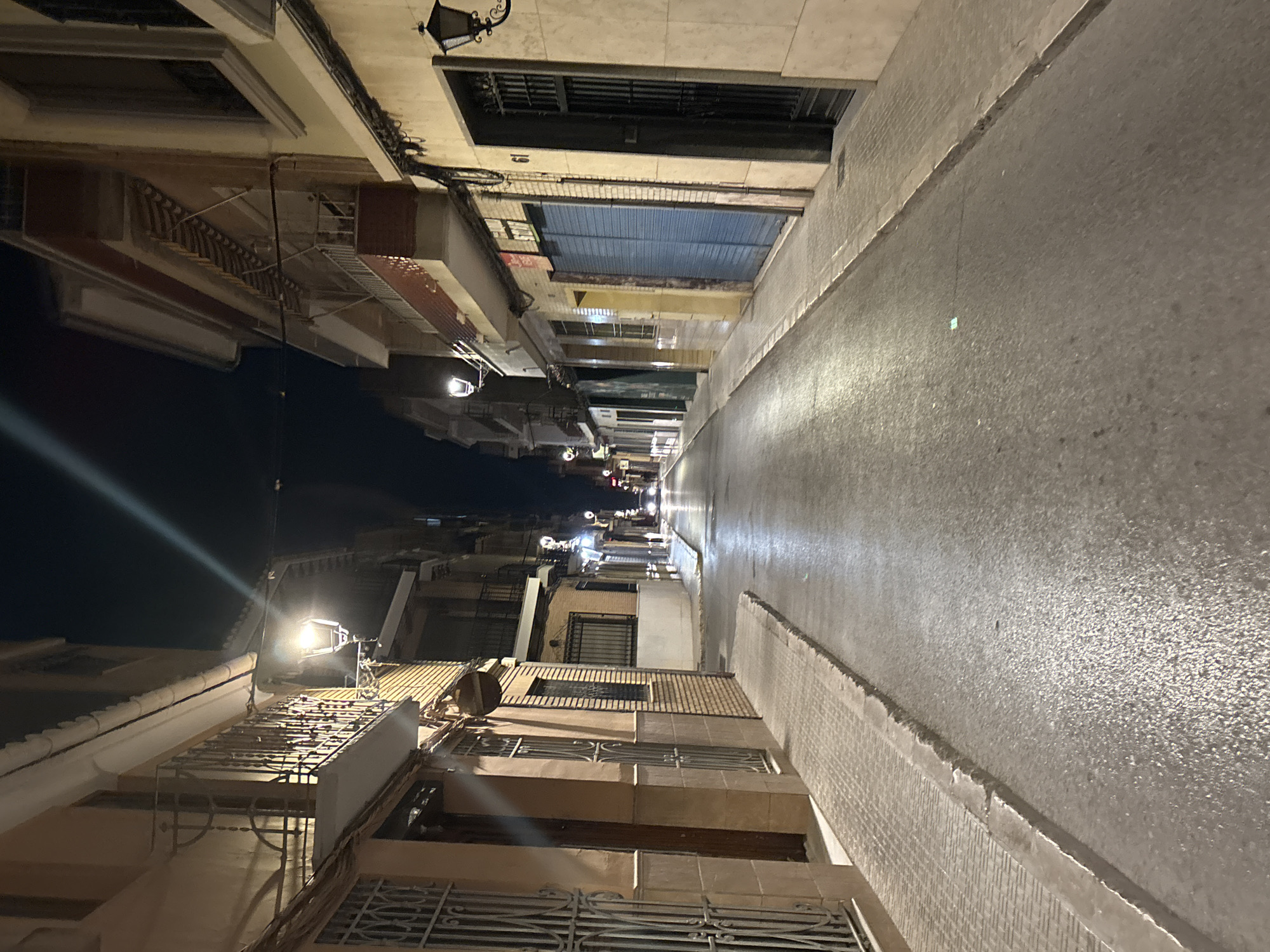 The image depicts a street scene at night in Picassent, Spain. The perspective is from street level, looking down a narrow street lined with multi-story buildings on both sides. The street is paved with asphalt and appears wet, reflecting the light from streetlamps and building lights.
On the left side of the street, buildings feature balconies with wrought-iron railings and windows with security bars. The building facades are a mix of light brown and brick. There are doorways and what appear to be shop fronts with metal shutters. A sidewalk runs along the left side of the street, made of paving stones with a rough texture.
On the right side of the street, buildings have a lighter colored facade. There are windows, balconies, and doorways. One building has a prominent blue rolling shutter. A traditional black streetlamp is mounted on the wall of a building, to the right of the blue shutter. Another streetlamp is visible further down the street, on the left side.
In the distance, at the end of the street, car headlights are visible, suggesting ongoing traffic. The sky is dark, indicating it is nighttime. The overall impression is of a quiet urban street after dark.