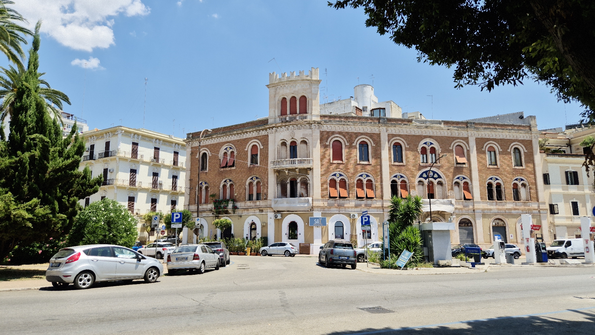 The image depicts a sunny day scene on a street in Taranto, Italy. The focal point is a large, multi-story building with ornate brickwork and arched windows. To the left of this building is a lighter-colored, more modern building. Several cars are parked on the side of the street and in a parking area in front of the buildings. Green trees and palm trees are also visible, adding to the urban landscape. The sky is clear blue with a few wispy clouds. The overall impression is that of a well-established urban area on a pleasant day. Specifically, the building in the center of the image is the Palazzo Stabile, located on Via Pupino in Taranto.