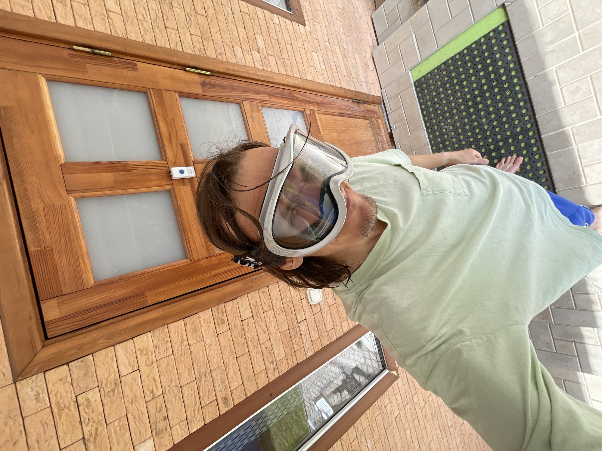 A man wearing large, white-framed goggles is indoors, near a wooden-framed window with a tan brick exterior. The man is wearing a light green shirt and blue pants. His right foot is visible on a black and green rubber mat that lies on tan tile flooring. A wooden door is visible in the background. The location is Перевозное, Russia.