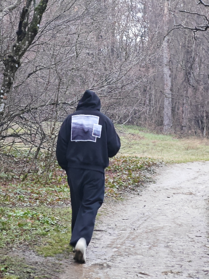 The image displays an individual walking away from the camera on a wet, unpaved dirt path through a forested area, consistent with the Shumen Plateau Nature Park in Shumen, Bulgaria. The subject wears a dark hooded sweatshirt, with the hood up, obscuring the head. The back of the sweatshirt features a large rectangular graphic depicting a dark, blurry landscape, overlaid by a smaller, lighter rectangular image on its lower right side. Dark trousers and light-colored shoes, which appear wet or muddy, are also worn. The individual's right foot is partially blurred, indicating forward motion. The path is damp with visible moisture. To the left, a mature tree with bare, moss-covered branches stands amidst sparse green ground cover and fallen leaves. The background consists of a dense stand of bare deciduous trees under an overcast sky, characteristic of a dormant season.