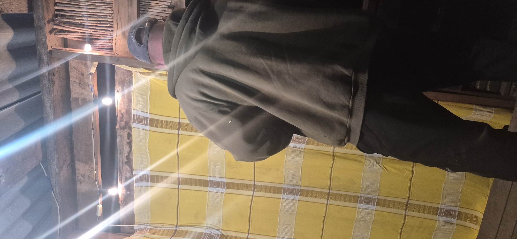 A male individual is depicted from the rear within an interior space in the city of Unknown, Unknown, characterized by rustic and functional construction. The individual wears a grey baseball cap with "PEPE JEANS" inscribed on the back in white lettering, an olive-drab hooded sweatshirt with the hood down, and dark, possibly black, casual trousers.
The overhead structure comprises corrugated metal roofing, supported by rough-hewn wooden beams. Exposed electrical wiring is visible, strung between beams and connected to two bare incandescent light bulbs, which emit strong light and cause prominent lens flare across the image. To the left of the individual is a yellow fabric, possibly a curtain or tapestry, featuring a grid pattern of white and brown stripes, with some faint darker marks visible. On the right, a vertical arrangement of thin wooden sticks or branches forms part of a wall or partition, adjacent to a darker wooden structure partially visible at the bottom right. The floor beneath the individual is dark and indistinct. The person is standing, facing away from the camera.