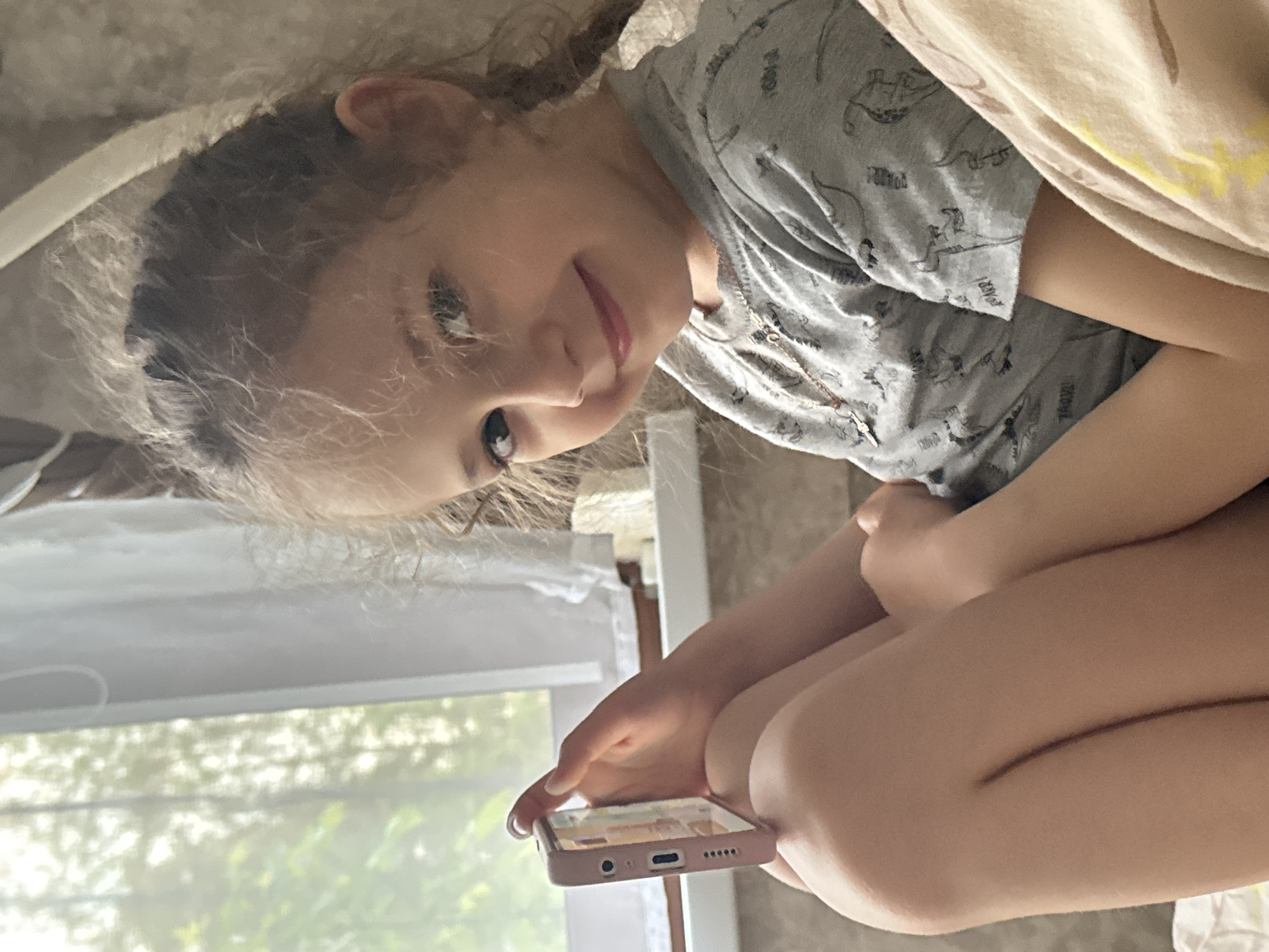 A young girl with fair skin and light brown hair is sitting indoors, looking down at a smartphone held in her hands. She wears a gray t-shirt with dinosaur patterns. She is positioned near a window that shows green foliage outside. The image is taken at an unusual angle, suggesting the viewer is positioned higher than the subject. Her legs are drawn up towards her chest. The location is Раздолье, Russia.