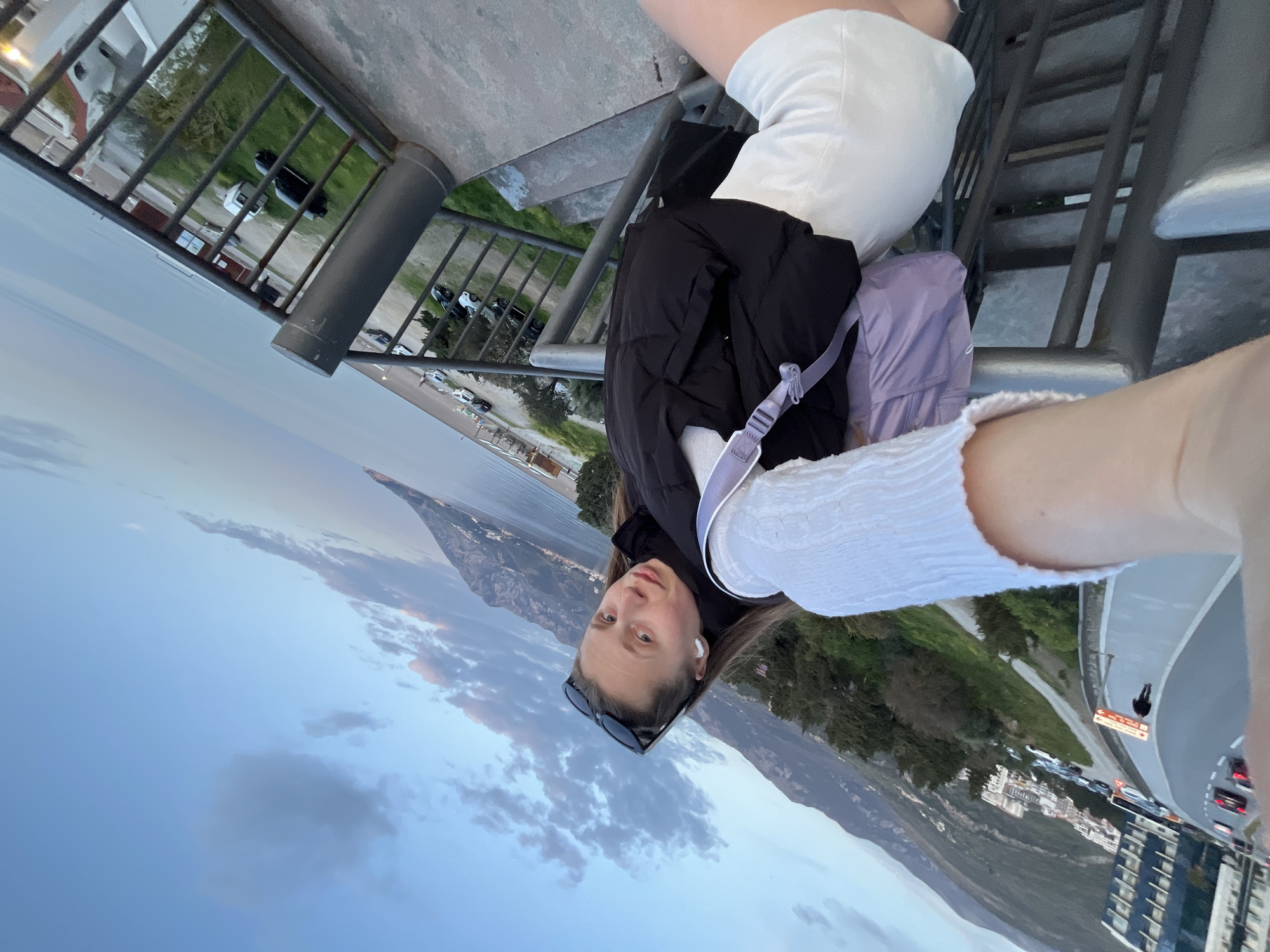 The image is a selfie taken on a structure with metal railings and stairs. The person taking the selfie is wearing white shorts, a white long-sleeved shirt, and a black jacket, and has a purple backpack. They are also wearing sunglasses and have an earbud in their ear.
The background shows a cityscape with buildings and a winding road with cars. In the distance, there is a mountain range and the sea. The sky is partly cloudy with shades of blue and grey.
Based on the landscape and architecture, the location appears to be Boreti, Montenegro.