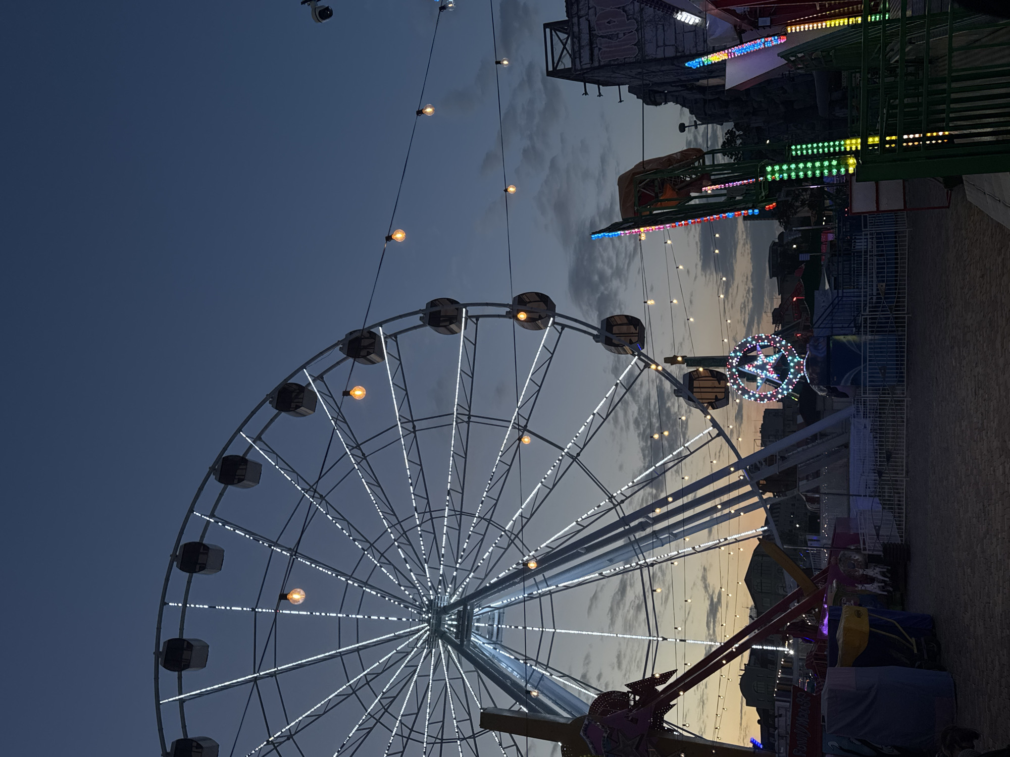 The image depicts a Ferris wheel at night in Yevpatoriya, Ukraine. The wheel is lit with white lights and has dark passenger cars. Additional carnival rides and structures are visible in the background, illuminated with various colored lights. Above the rides, the sky transitions from a darker blue to a lighter sunset hue. Strings of lights hang above the scene. The image is vertically oriented.