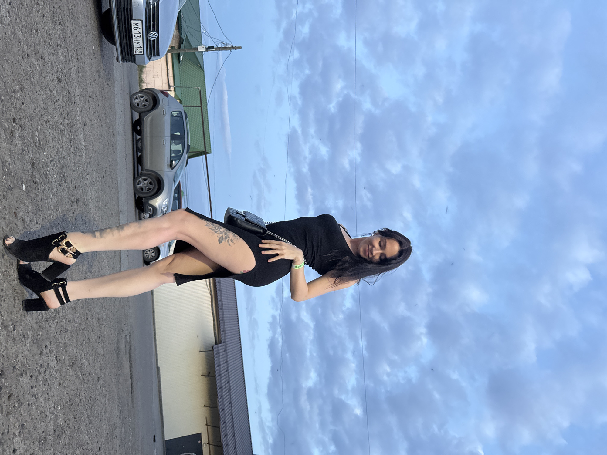 A woman stands on an asphalt street in what appears to be an urban environment. She is wearing a short black dress, black open-toe high heels, and a small black shoulder bag. She has dark hair and a light complexion. Her pose suggests she is looking down at her stomach. Visible on her leg is a tattoo. Behind her is a metal-sided building with a peaked roof. In the background are two light-colored cars, one of which has a Russian license plate reading "M613HY 159". The sky is overcast with light blue and grey clouds. The location is likely Chaikovsky, Russia, based on the license plate information.