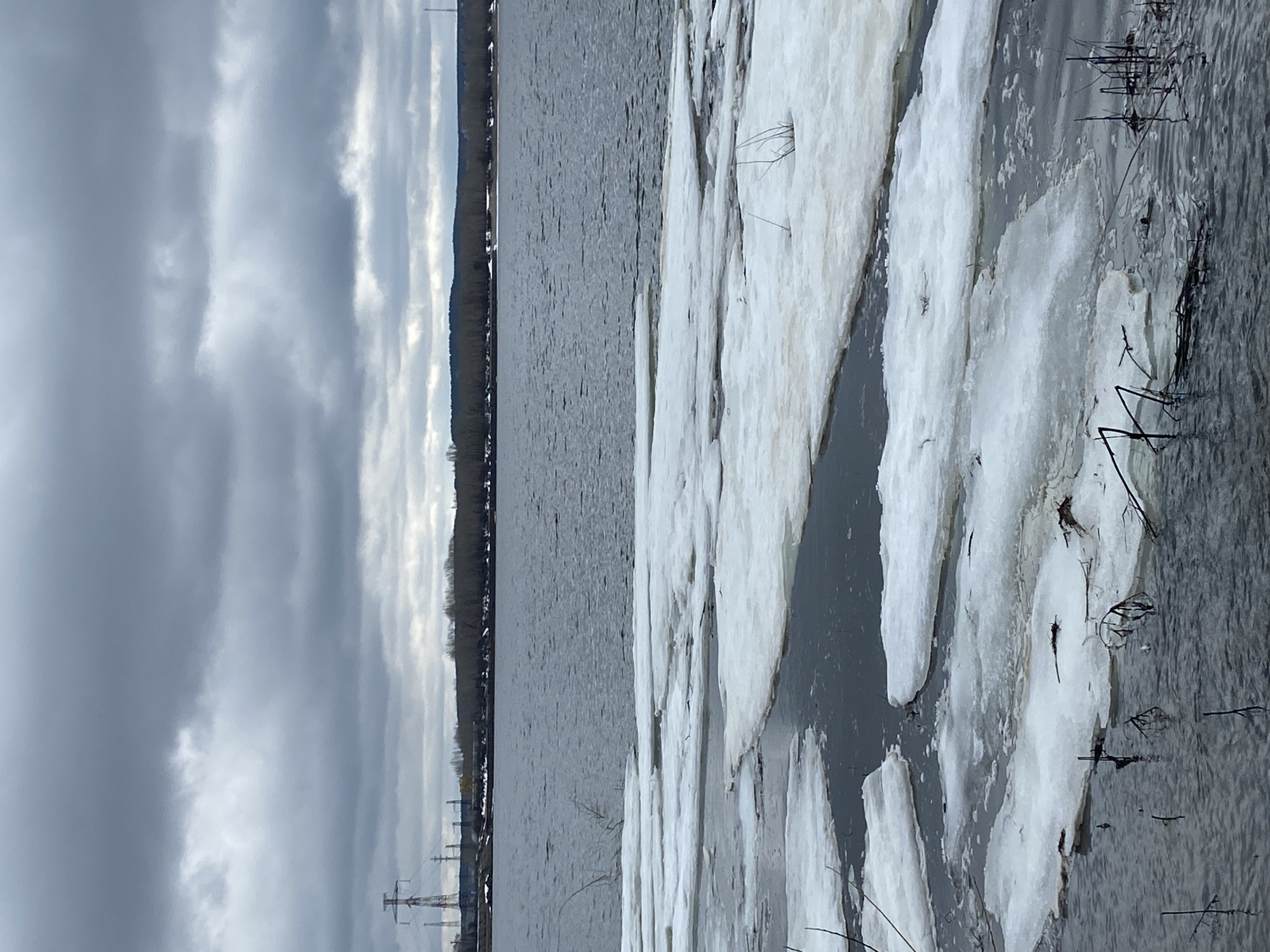 The image depicts a body of water, likely the Kama River, with ice floes accumulating along the shore. The sky is overcast with heavy clouds. In the distance, a treeline marks the opposite bank of the river. Power lines are visible on the near shore. The location is consistent with Chaikovsky, Russia, given the presence of the Kama River and the typical winter/spring conditions of ice breakup. No people or animals are visible in the image.