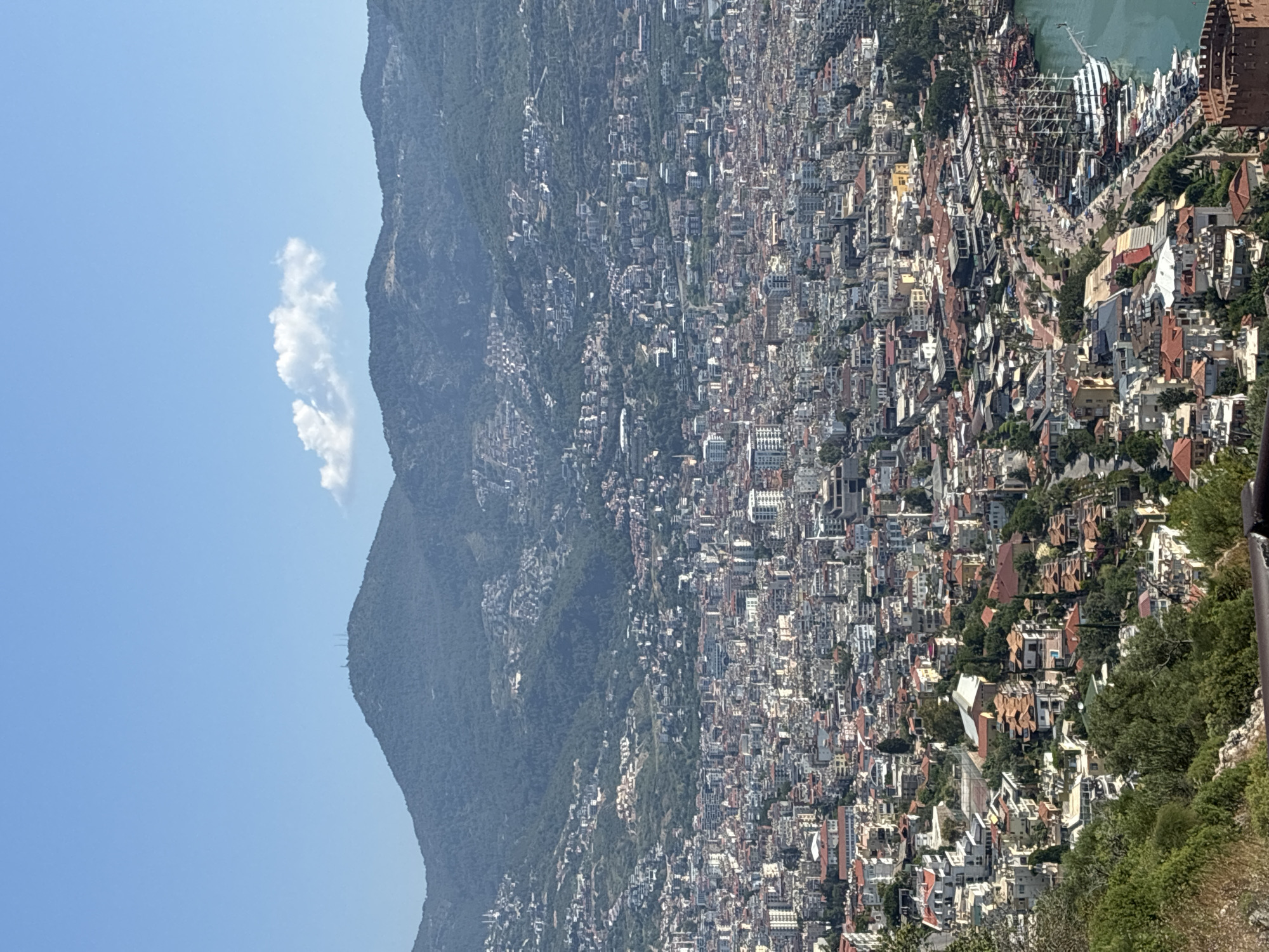 The image shows a panoramic view of Alanya, Turkey, from a high vantage point. In the foreground, lush green trees and rocky terrain descend towards the city. The city itself is densely packed with buildings of varying heights and architectural styles, many featuring red-tiled roofs. The urban landscape extends towards the coast, where a harbor filled with numerous boats and ships, including several large, traditional sailing vessels, is visible. Beyond the city, a range of majestic mountains forms the backdrop, with one peak topped by communication towers. The sky above is a clear, bright blue with a single, wispy white cloud. The overall scene captures the vibrant coastal city nestled against a dramatic mountainous terrain.
