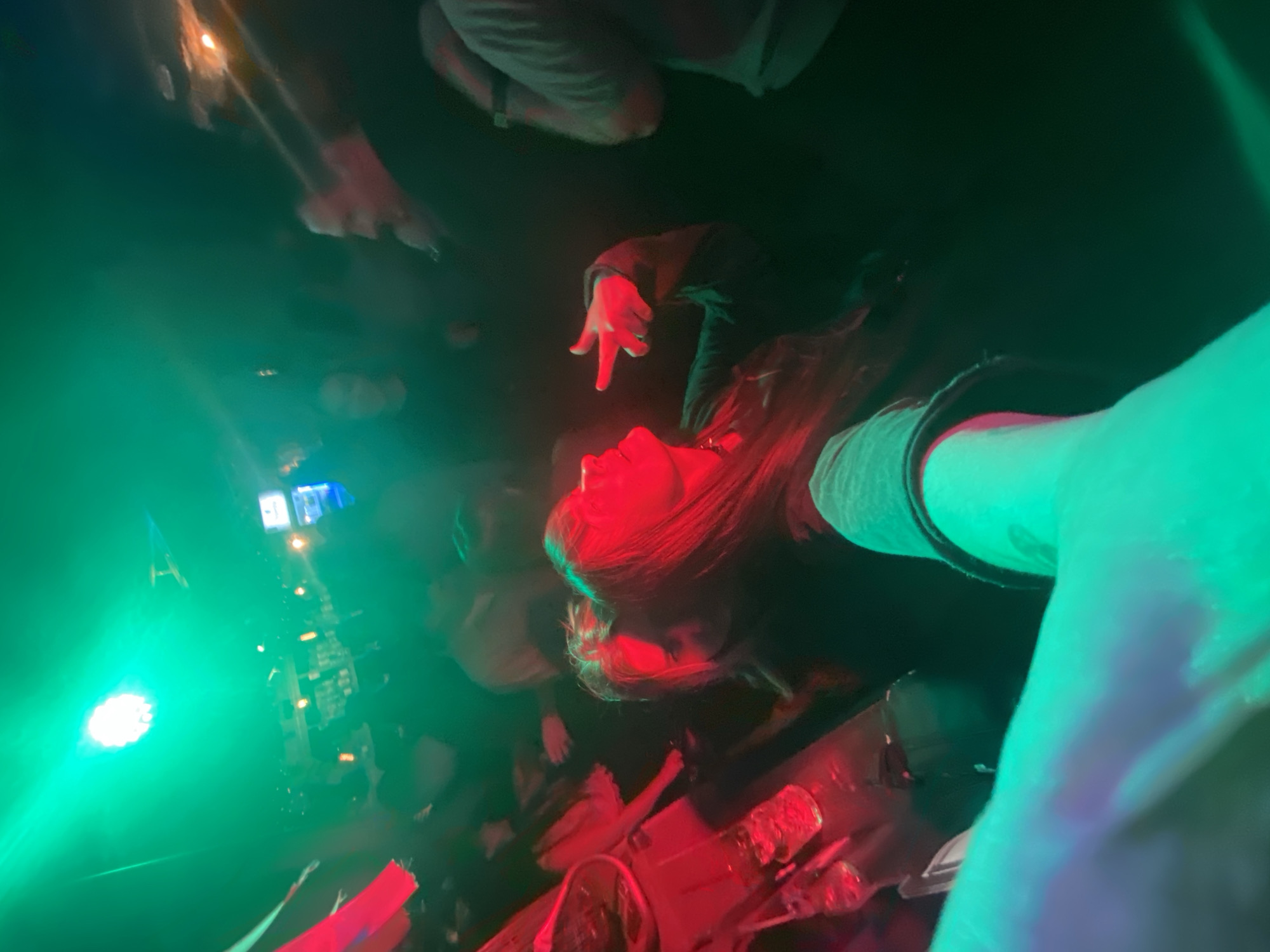 The image shows an indoor scene with multiple people, predominantly illuminated by green and red stage lighting. A person with red hair, wearing a dark jacket, is looking up with their mouth slightly open. Above them, another person's hand is extended downward, seemingly pointing towards the person below. Other individuals and objects are visible in the background, but are partially obscured by the lighting and depth of field. A bright green light source is prominent on the left side of the frame. Red light highlights the lower portion of the scene. The city of Izhevsk, Russia is not visible or discernible from the image.