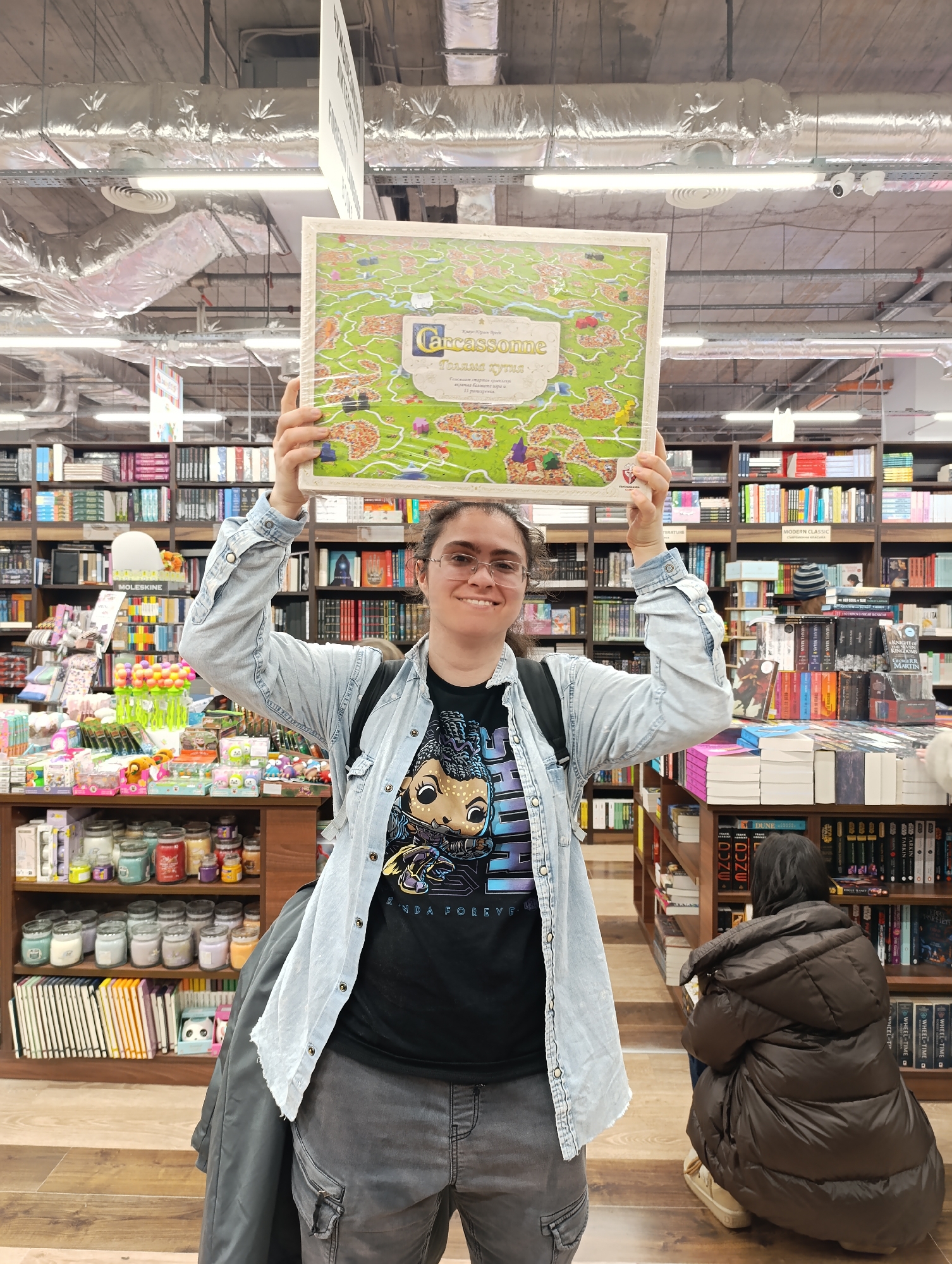 Here is a detailed description of the image:
**Scene Overview:**
The image appears to be taken inside a bookstore in Sofia, Bulgaria. The woman is standing in an aisle filled with bookshelves on both sides. Another person is kneeling and looking at the books in the background.
**People:**
The main subject is a woman holding a board game above her head. She is wearing glasses, a black t-shirt with a graphic design, a light blue denim jacket, and grey cargo pants. She has a backpack on. She is smiling at the camera. Another person is kneeling in the background, facing the bookshelf, wearing a brown jacket.
**Objects:**
The most prominent object is the board game "Carcassonne", which the woman is holding up. The bookstore has rows of shelves filled with books. There is also a display table with various items, including candles, notebooks, and toys.
**Environment:**
The store has a warehouse-like feel with exposed ceiling pipes and metal beams. The bookshelves are packed with a variety of books. The lighting is bright and somewhat artificial. There is a display table with candles and toys on the left.
**Location:**
Based on the Cyrillic text on the game box ("Голяма кутия"), the location is likely Sofia, Bulgaria.