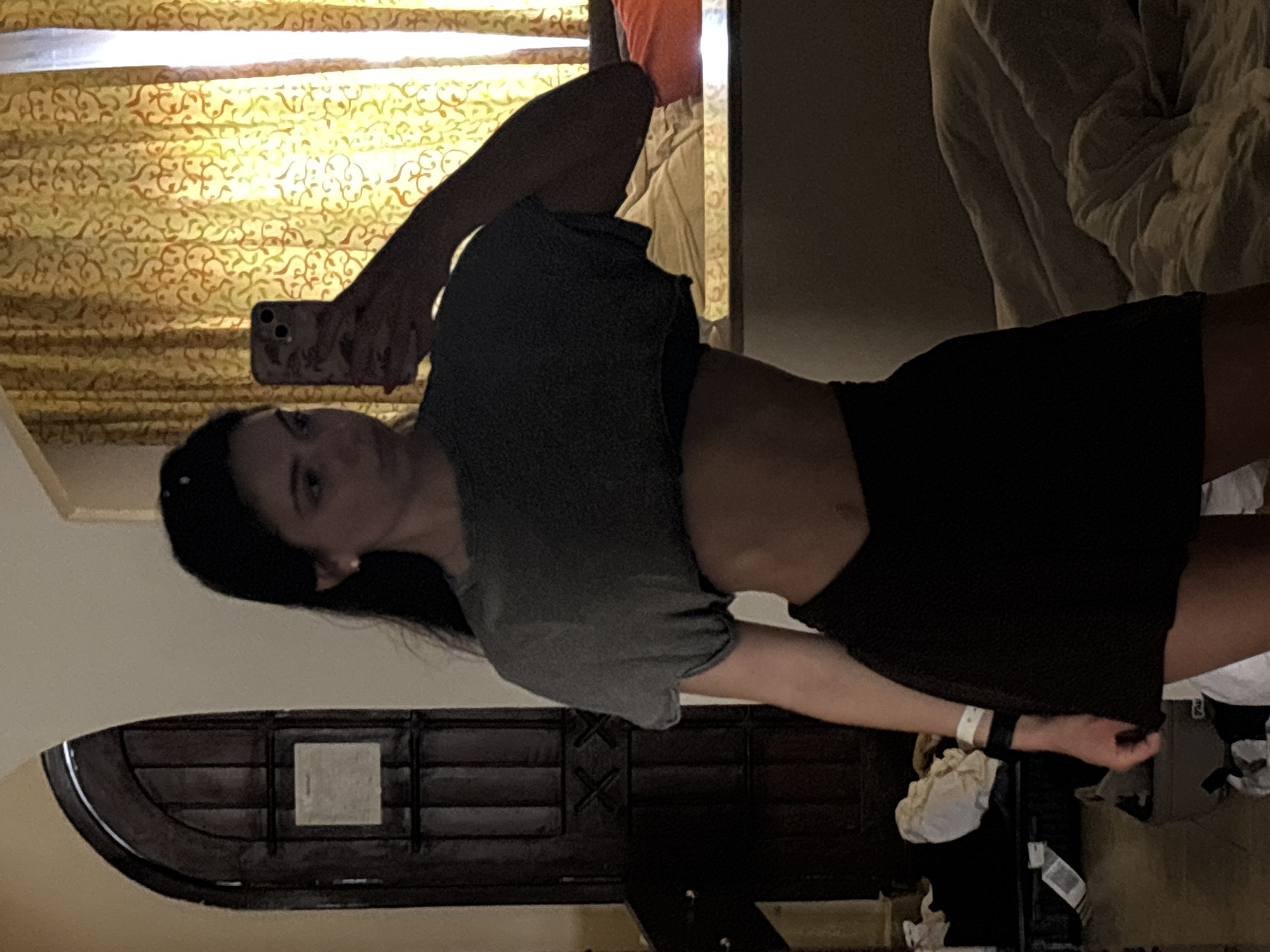 A woman is taking a selfie in a room in an unknown location in the city of Unknown, Unknown. She is wearing a gray crop top and a black skirt. Her left hand holds a smartphone. The background includes a patterned curtain, a bed with pillows, a doorway, and miscellaneous items on the floor.