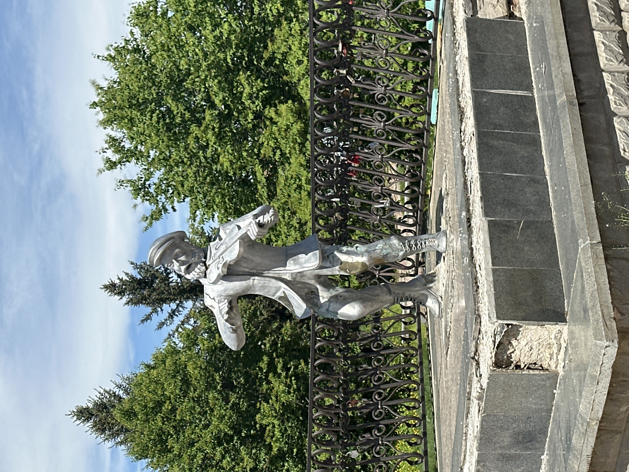 A bronze statue of a man in historical clothing stands next to an ornate black iron fence. The statue has boots and a hat and appears to be in mid-stride. The statue is positioned next to a set of grey stone steps or a monument base. Lush green trees are in the background, partially obscuring a blue sky with clouds. The statue is located in Birobidzhan, Russia.