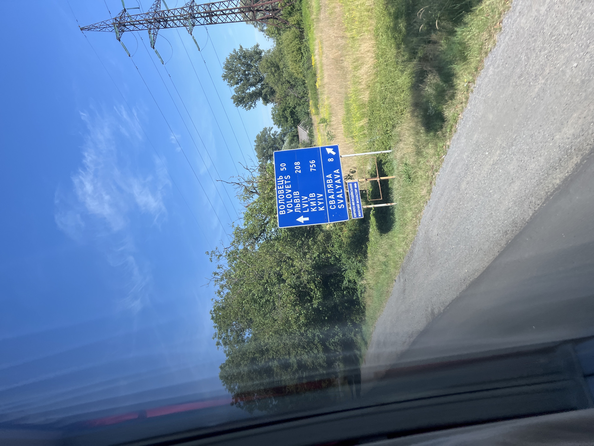 The image shows a road sign in Suskovo, Ukraine, indicating directions to Volovets (50 km), Lviv (208 km), and Kyiv (756 km). It also shows a direction to Svalyava (8 km) with an arrow pointing right. A smaller sign below indicates a monastery at a distance. The signpost is located in a rural area with green trees and fields. A power line tower is visible in the background, along with a blue sky with scattered clouds. The perspective suggests the photo was taken from a moving vehicle.