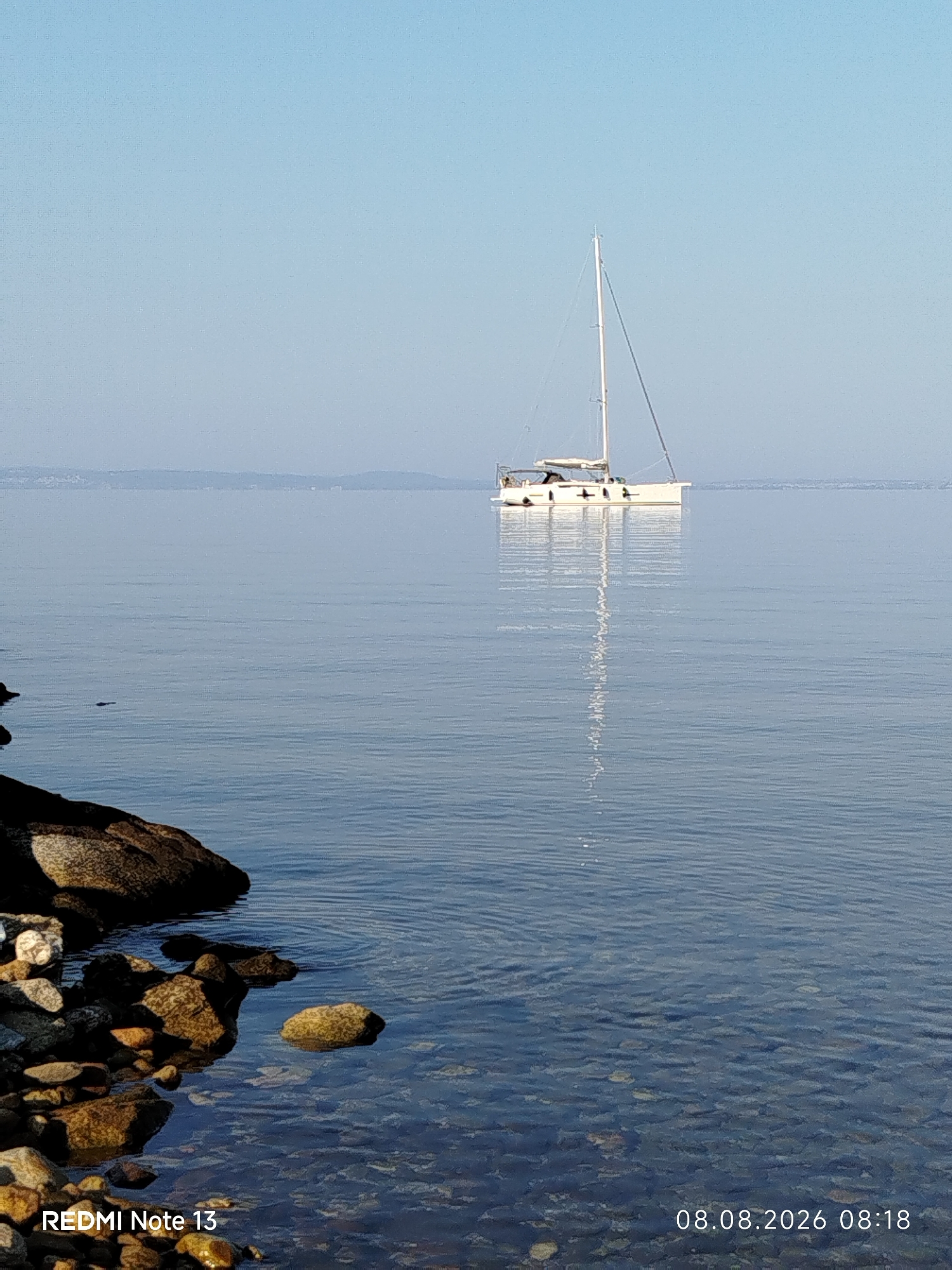 A still, clear body of water with a sailboat anchored in the distance, a rocky shoreline in the foreground, and a hazy coastline in the background. The sailboat is white with a tall mast. The water is calm, reflecting the sky and the sailboat. The shoreline is composed of various-sized rocks and pebbles. The sky is clear and blue. The image has a timestamp in the bottom right corner: "08.08.2026 08:18". There is also text in the bottom left corner that reads "REDMI Note 13", indicating the device used to capture the photo.