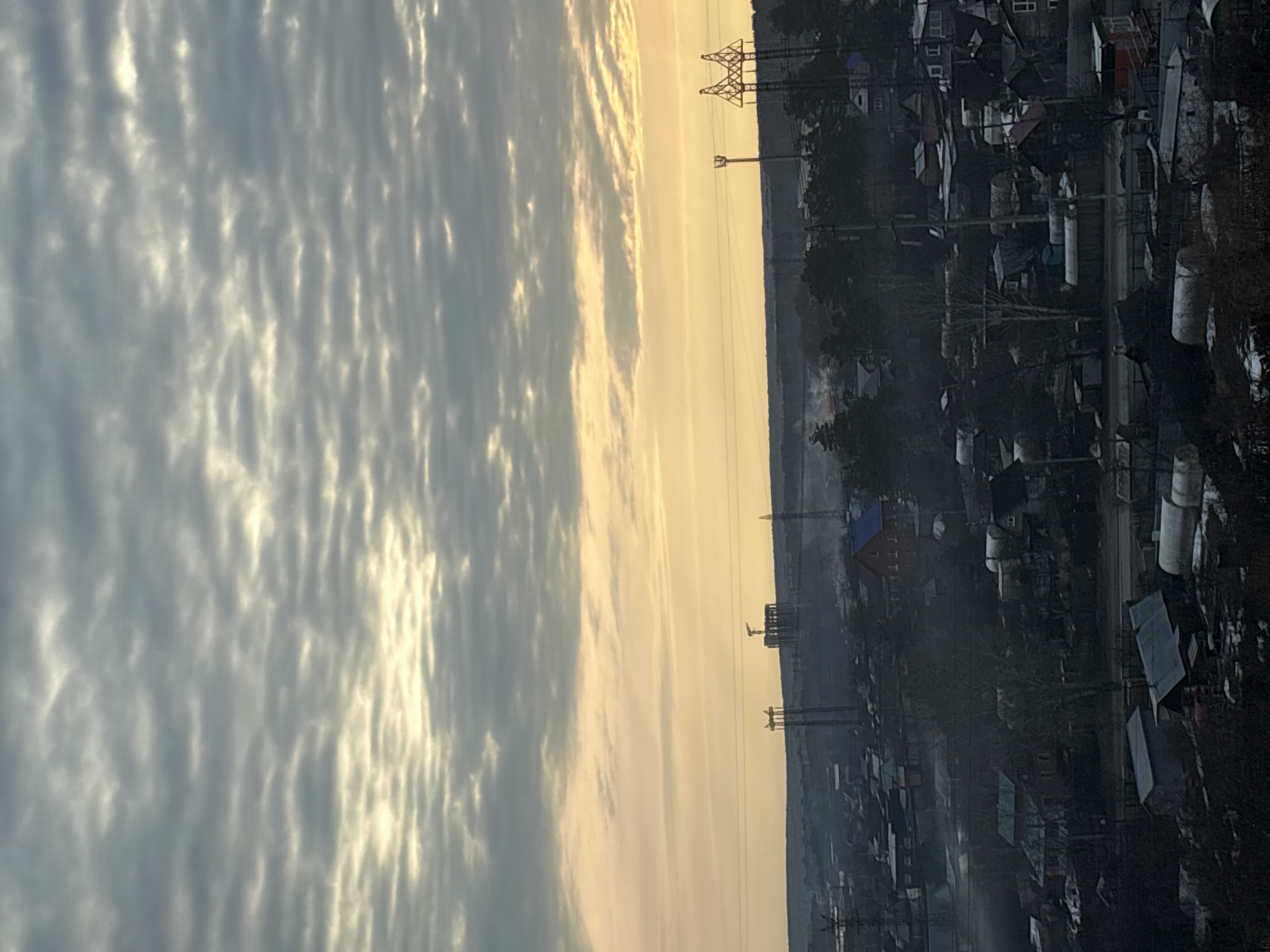 The image shows a cityscape in Chaikovsky, Russia, at dawn or dusk. The sky occupies the left half of the image and is filled with patterned clouds, tinged with golden sunlight. On the right side of the image, a built-up area with houses, buildings, and infrastructure is visible. Electrical pylons and lines cut through the scene. The overall atmosphere is hazy, and the lighting suggests it may be early morning or late evening.
