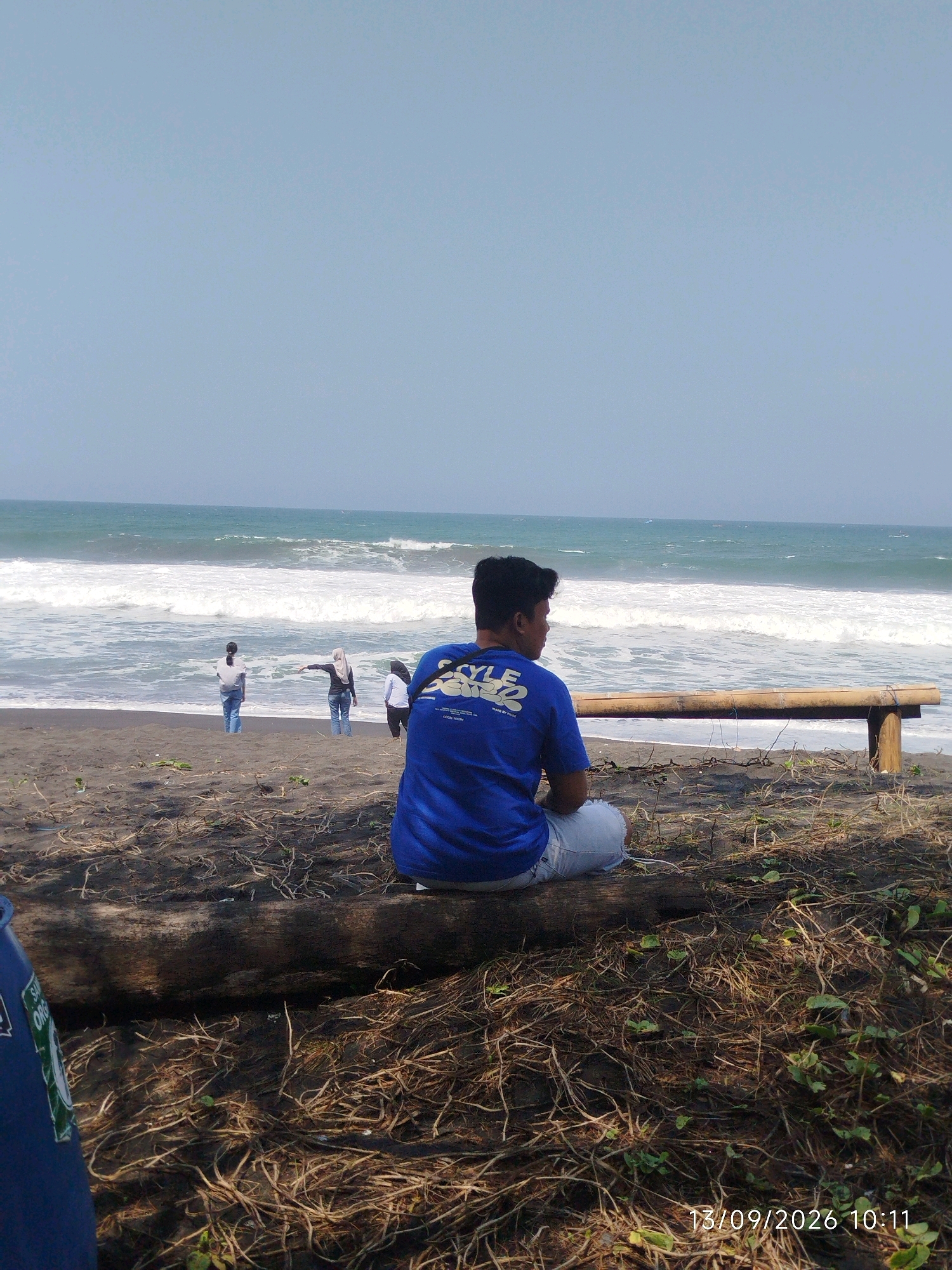 A young man in a blue t-shirt and light-colored shorts is sitting on a log on a sandy beach, looking out at the ocean. The waves are crashing on the shore. In the background, there are three other people standing near the water. The sky is clear and blue. There is a wooden bench to the right of the man. The scene appears to be in Pleret, Indonesia. The timestamp on the image indicates it was taken on September 13, 2026, at 10:11 AM.