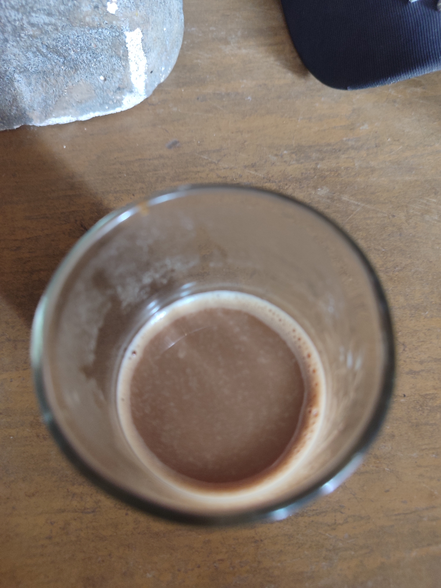 A close-up, overhead shot of a glass filled with a brown beverage sits on a wooden surface. The beverage has a light froth around the rim. To the left, a portion of a light-colored, rough-textured object is visible, and to the top right, a dark blue object, possibly a hat, is partially in frame. The scene is illuminated by overhead lighting, creating reflections on the glass and beverage. The location is Gantungan, Indonesia.