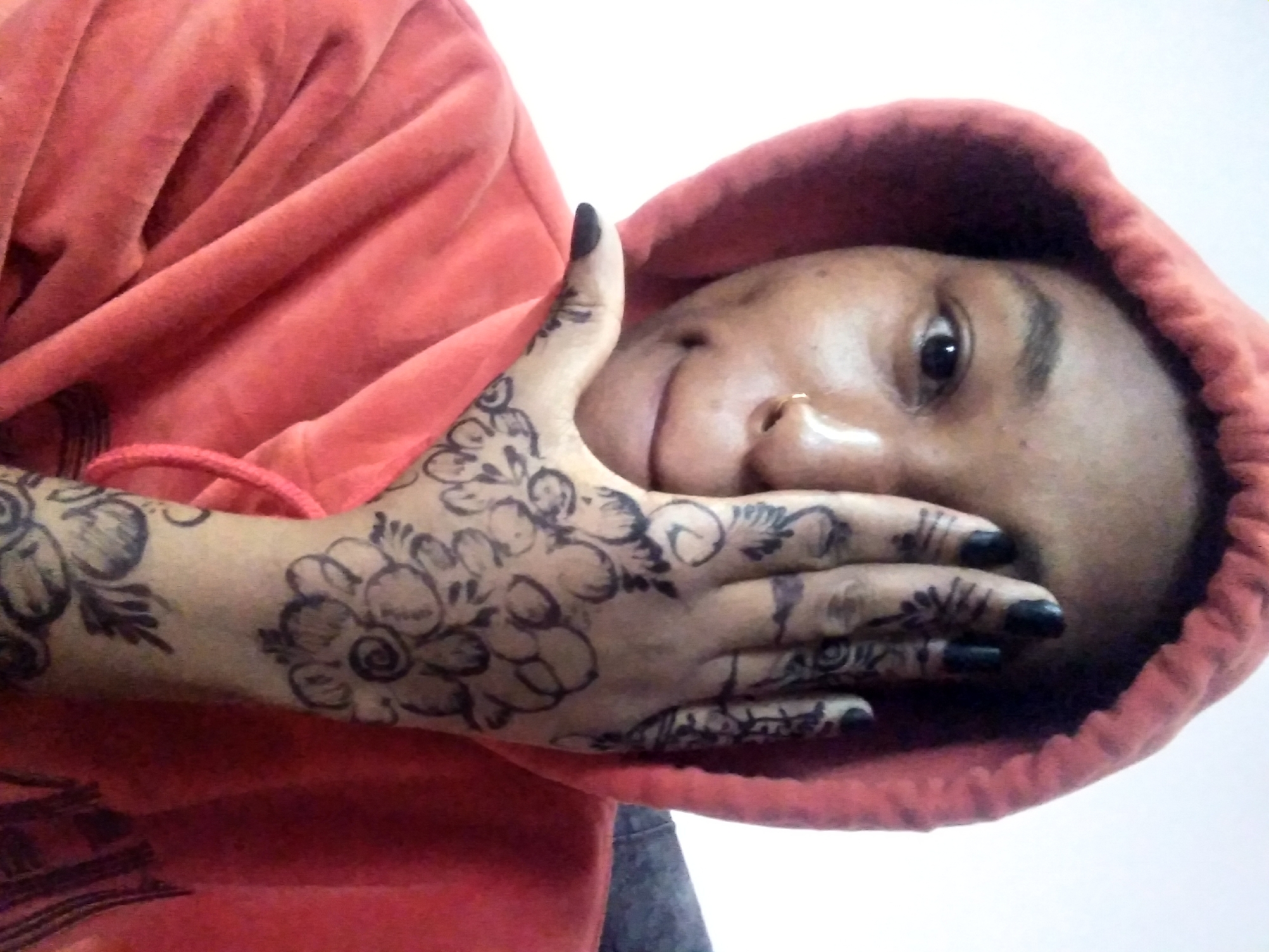 A close-up studio shot features a person wearing an orange hoodie, with the hood pulled up, partially obscuring their face. The person's face, featuring a nose ring on the left nostril, is positioned to the right side of the frame. Their right hand, adorned with henna tattoos of floral designs, is raised, partially covering the face. The fingernails are painted black. The background is a plain white wall.