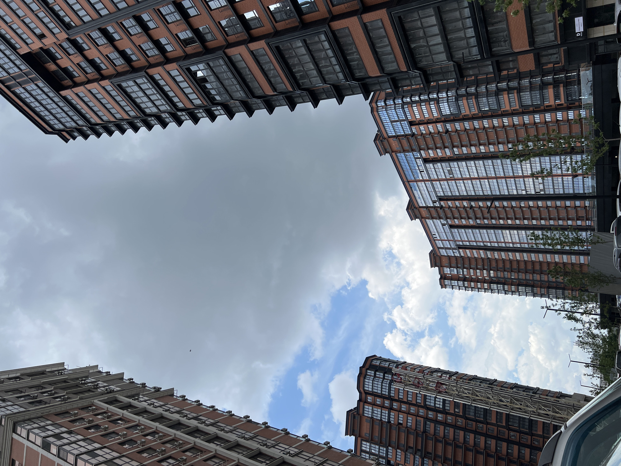 In Krasnodar, Russia, three modern, multi-story residential buildings with red brick facades and dark-framed windows are captured from a low-angle perspective. The buildings feature multiple balconies and a contemporary architectural style. The sky above is a mix of dramatic grey clouds and patches of blue sky dotted with white, fluffy clouds. In the foreground, the roof and windshield of a silver car are visible, suggesting an urban street or parking area. The environment appears to be a developed residential district, with streetlights and some greenery visible at the base of the buildings.