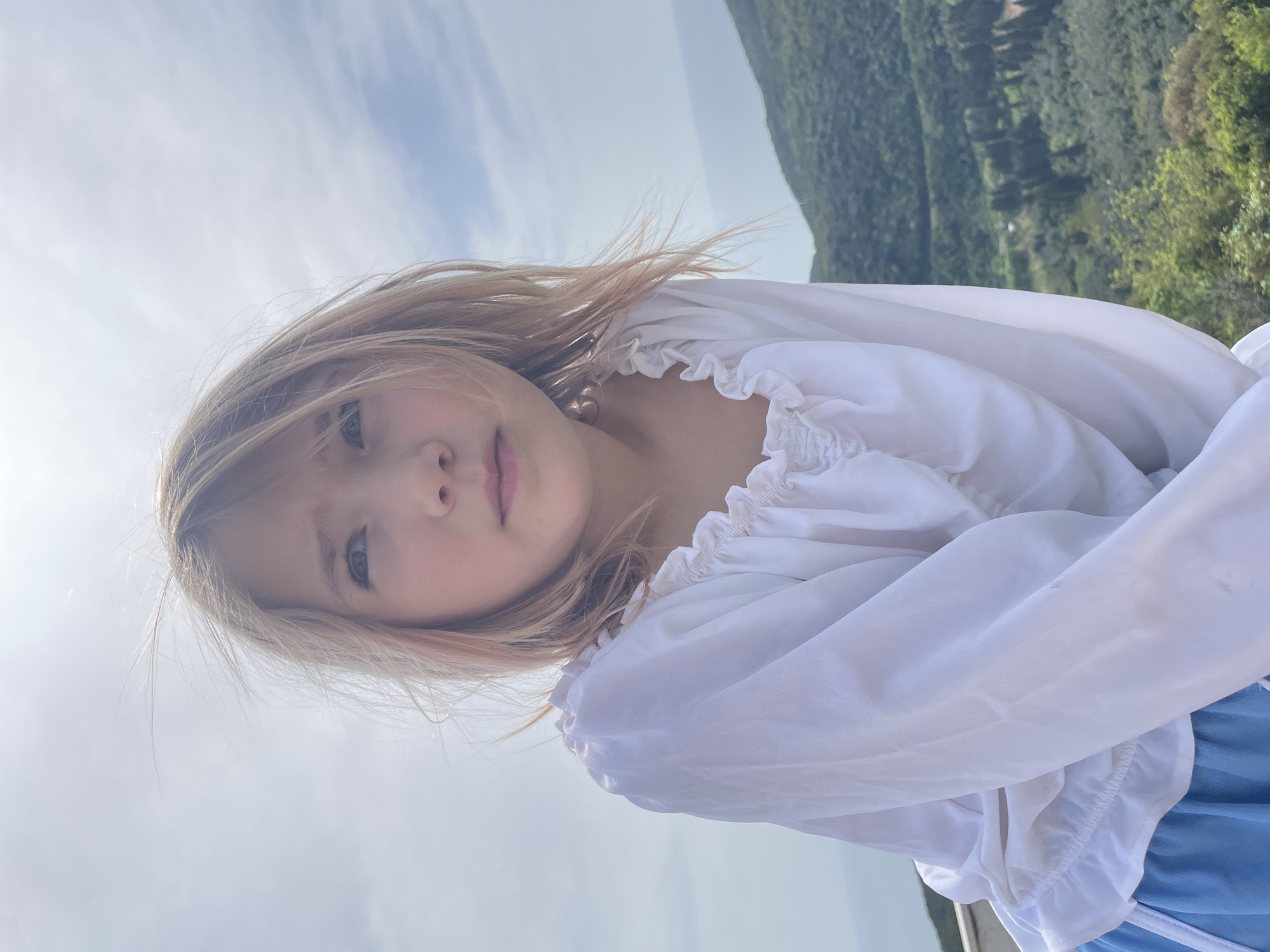 A young girl with long, light blonde hair and light-colored eyes is featured in a vertically oriented portrait, looking directly at the viewer with a calm, neutral expression. She wears a white top with ruffled details around the neckline and elasticated sleeves, and a hint of a blue garment, possibly a skirt, is visible towards the bottom right of the frame. Her hair is slightly windswept.
The background is split between a bright, partly cloudy sky on the left side (which would be above her if the image were rotated correctly) and a lush, verdant mountainous or hilly landscape on the right. The green hills are densely covered with trees and vegetation, stretching into the distance under the clear daylight.
The image appears to be taken outdoors during the daytime, under bright but soft natural light. The setting is a scenic natural environment, consistent with the provided location of Bratica - Braticë, Montenegro. No visible text is present in the image.