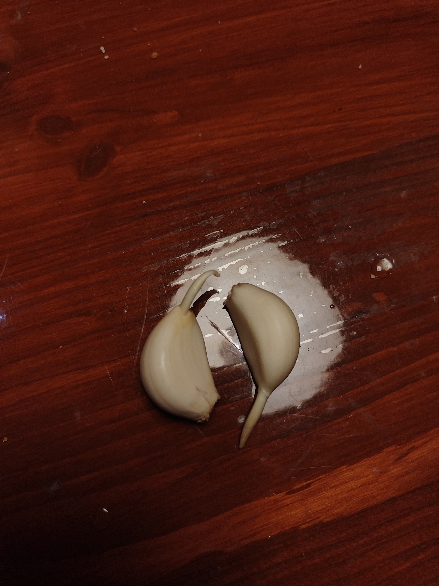 Two peeled garlic cloves are placed on a dark reddish-brown wooden surface, possibly a table or countertop. The wood displays a visible grain and appears to be varnished or polished. One clove is positioned on the left, slightly curved with a defined root end pointing upwards. The second clove is to its right, also curved, and rests with its root end pointing downwards and slightly towards the right.
Beneath and around the garlic cloves, there is a wet-looking, somewhat opaque residue or spill on the wooden surface, forming an irregular, shiny patch. This liquid appears to have dried in some areas or is semi-translucent, showing streaks and small droplets. Scattered on the wooden surface, particularly in the upper half and edges of the image, are a few small, light-colored specks, possibly crumbs or salt crystals. The lighting suggests an indoor setting. There is no visible text, and no specific activity or emotions are depicted. The time of day and weather cannot be determined.