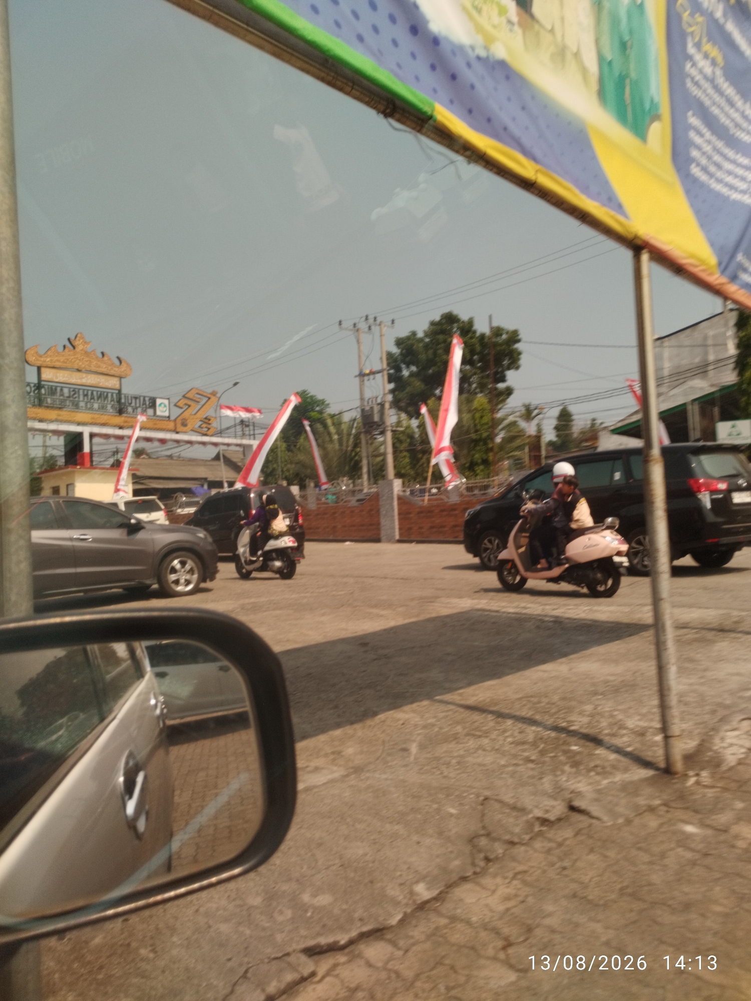 This image captures a street scene in Bandar Lampung, Indonesia, with a prominent banner featuring the words "PEMERINTAH KOTA BANDAR LAMPUNG" and what appears to be a logo with the number "75." The banner is supported by poles and hangs over a street or parking area. In the foreground, a gray car's side mirror reflects the surrounding environment, showing the car's door handle and the concrete ground.
Further into the scene, several vehicles are present. A silver Honda HR-V is parked on the left. To its right, a dark-colored SUV and a black minivan are visible. A person wearing a helmet and a beige jacket is riding a light pink scooter, which is positioned between the SUV and minivan. Another scooter, possibly a Yamaha Mio, is seen to the left of the person on the pink scooter, with a rider in a black outfit.
The background reveals a building with a red and yellow facade and a decorative golden archway, possibly an entrance. Red and white flags, characteristic of Indonesia, are displayed on poles throughout the area. The ground is paved with concrete. Power lines and utility poles are also visible against the hazy sky. The timestamp "13/08/2026 14:13" is overlaid on the bottom right corner of the image.