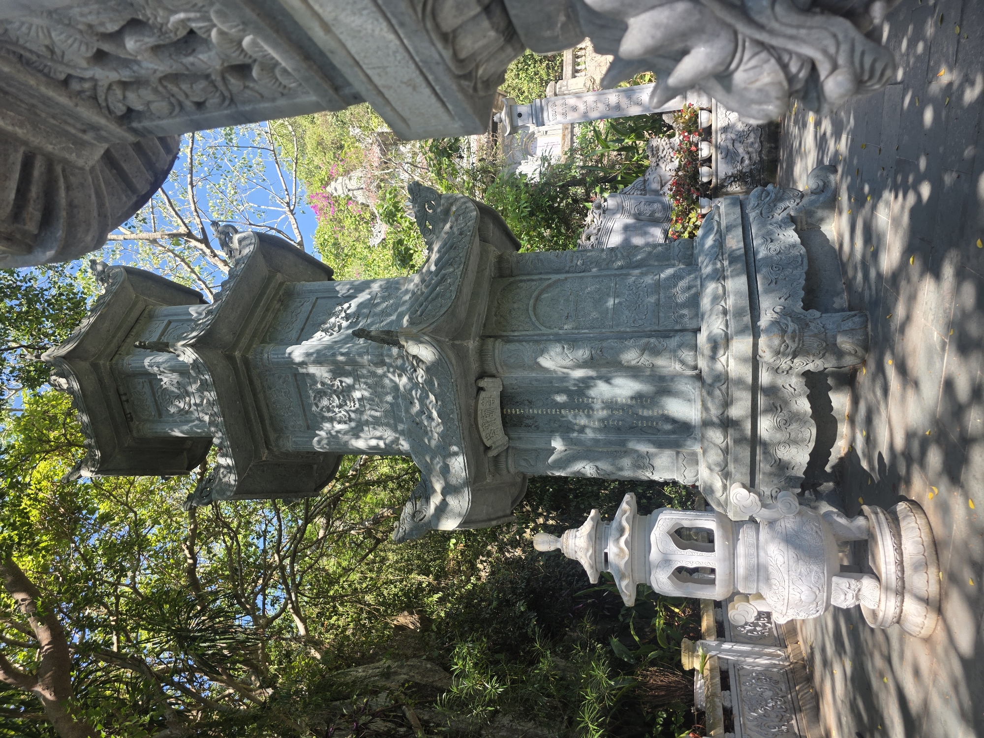 The image, rotated 90 degrees clockwise, depicts an outdoor scene at a temple or pagoda in Đà Nẵng, Vietnam, under a bright, sunny sky. The central focus is a towering, multi-tiered stone pagoda or stupa, intricately carved from grey stone. Each tier features detailed architectural elements and ornamental patterns, with some sections depicting what appear to be dragons or mythical creatures. Strong sunlight illuminates parts of the structure, while deep shadows highlight its sculptural depth.
To the right of the large pagoda, a smaller, white stone lantern-like ornament stands on a carved pedestal. This lantern also features intricate designs, including cloud motifs and cut-out sections. The ground beneath is paved with stone, and numerous small yellow leaves are scattered across it. The paving is dappled with shadows cast by trees and the structures, indicating direct sunlight, likely mid-morning or afternoon.
The background is filled with lush green trees and dense foliage, suggesting a natural, garden-like setting. Patches of bright pink flowers are visible among the greenery on a distant hillside. A clear blue sky is visible between the tree canopies.
Several instances of visible text appear on the stone elements. On the large central pagoda, a rectangular panel near the top bears an inscription in what appears to be Chinese characters. Further in the background, a tall, vertical stone monument also features text, seemingly in Vietnamese script. Partially visible words from this inscription include "NGUYEN HUU...", "SAU...", "TANH OAI...", and "CHAU GIANG...". Another stone pillar in the distance also has faint inscriptions. The scene conveys a sense of historical reverence and serene natural beauty.