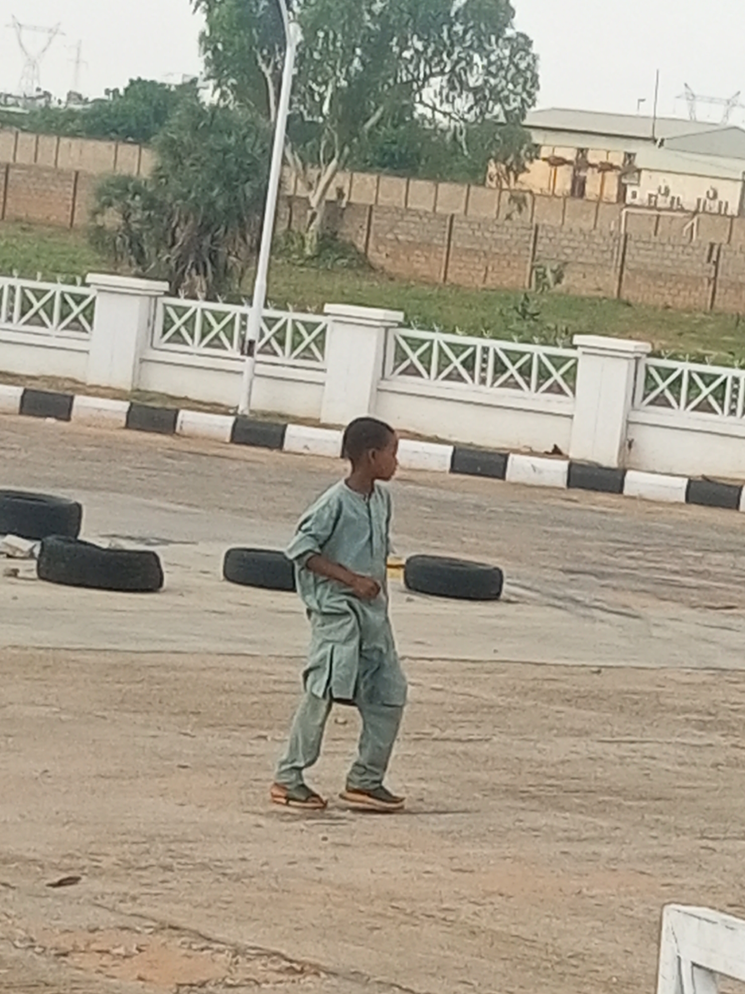 A young boy, wearing a light green traditional outfit and sandals, walks across a dusty, outdoor area. The boy is positioned in the lower right quadrant of the image, facing to his left. He appears to be walking casually.
The setting is an open, paved area, possibly a road or parking lot, with several discarded tires scattered on the ground. In the background, there is a white ornamental fence with a crisscross pattern. Beyond the fence, there is a grassy area with trees, and further back, buildings and electrical pylons are visible under an overcast sky. The lighting suggests it is daytime, likely late afternoon or early morning due to the soft, diffused light. There are no visible signs of text in the image. The overall scene is mundane, with no strong emotional indicators. The location context is Garko, Nigeria.