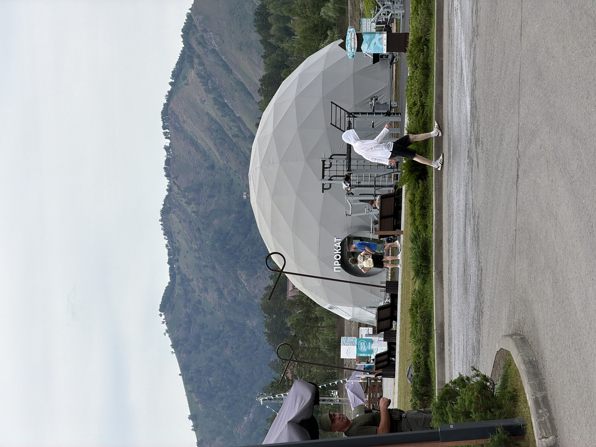 The image depicts an outdoor scene at a recreational facility with a large, geodesic dome structure. The dome, which appears to be a rental facility indicated by the Russian word "ПРОКАТ" (PROKAT) displayed on its side, is a prominent feature. In front of the dome, there are outdoor exercise equipment setups.
Several individuals are visible in the frame. A person wearing a white hooded jacket and black shorts is walking on a paved path in the foreground, moving from left to right. To the left of the dome, near a gray tent-like structure, a person in a green cap and green shirt is standing and looking towards the left, possibly a security guard or staff member. Two individuals are standing near the entrance of the dome, one wearing a light-colored backpack and the other in a blue t-shirt and shorts. Another person is partially visible sitting on a bench in front of the exercise equipment.
The environment includes a paved area, a grassy lawn with manicured bushes, and a backdrop of mountainous terrain covered with trees. The sky is overcast, suggesting a cloudy day. The overall scene suggests a public leisure or sports complex located in a natural setting, possibly in or near the city of Озерное (майминский), Russia, given the presence of Russian text.