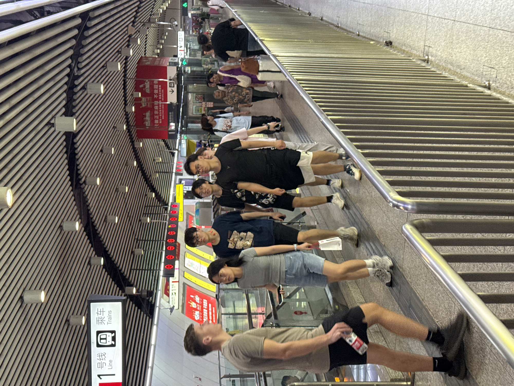 The image depicts a group of people standing in a line inside a subway station. The scene is identified as being located within the Yuzhong District, China, based on the signage visible in the image. The individuals in the line are diverse in age and attire. Several red and white posters, possibly advertisements or informational signs, are visible on the wall behind the group. There is a sign above the entrance with Chinese characters and the word "Trains" printed on it. A set of metallic stairs runs parallel to the line of people. The lighting in the station appears to be primarily provided by linear light fixtures installed along the ceiling.