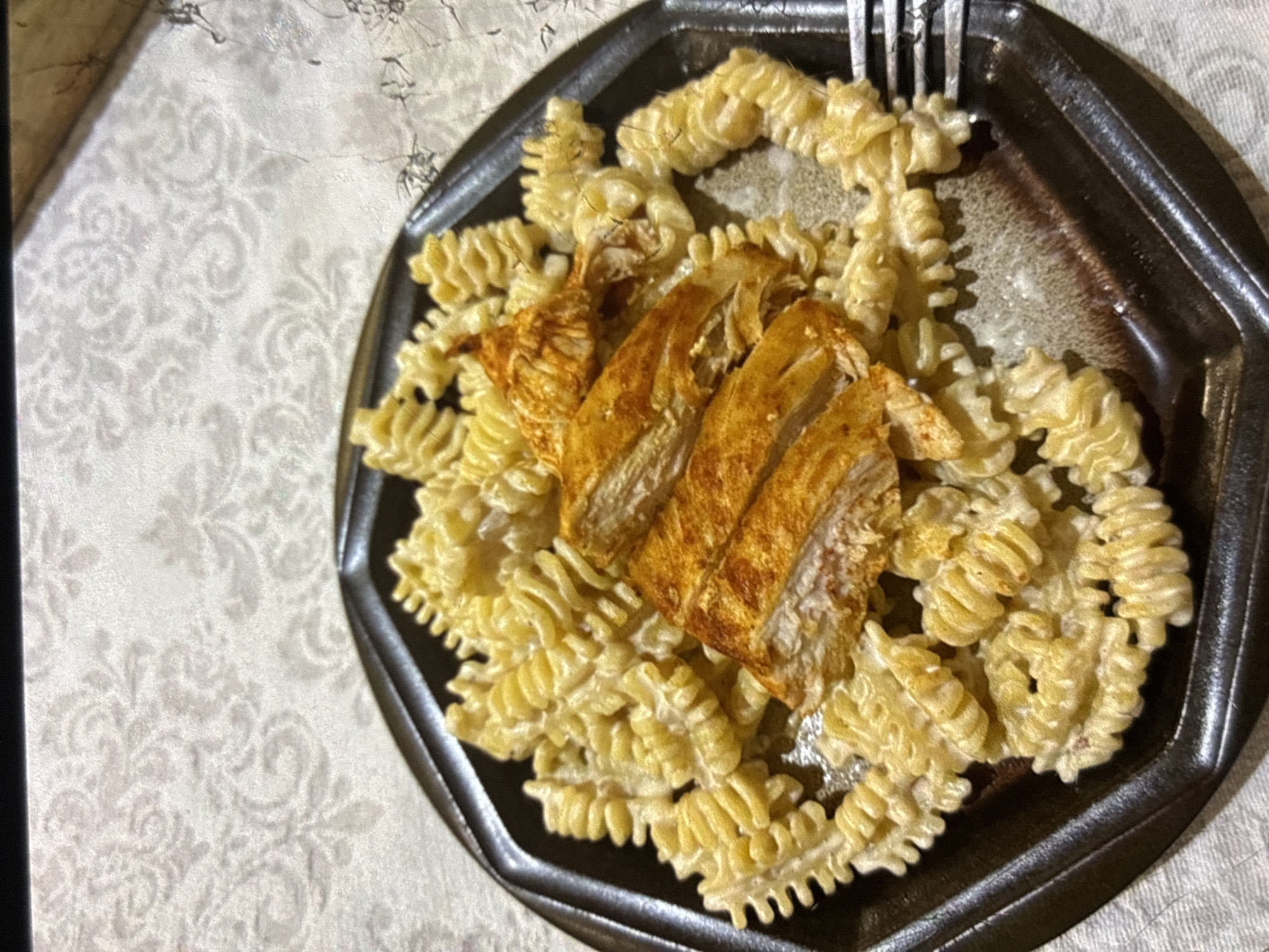 A plate of pasta and chicken sits on a patterned tablecloth. The pasta is rotini-shaped and covered in a creamy sauce. Sliced chicken breast, seasoned with what appears to be paprika, is placed on top of the pasta. A fork is partially visible on the right side of the plate. The background is out of focus, showing the textured surface of the tablecloth. This image was taken in Orihuela, Spain.