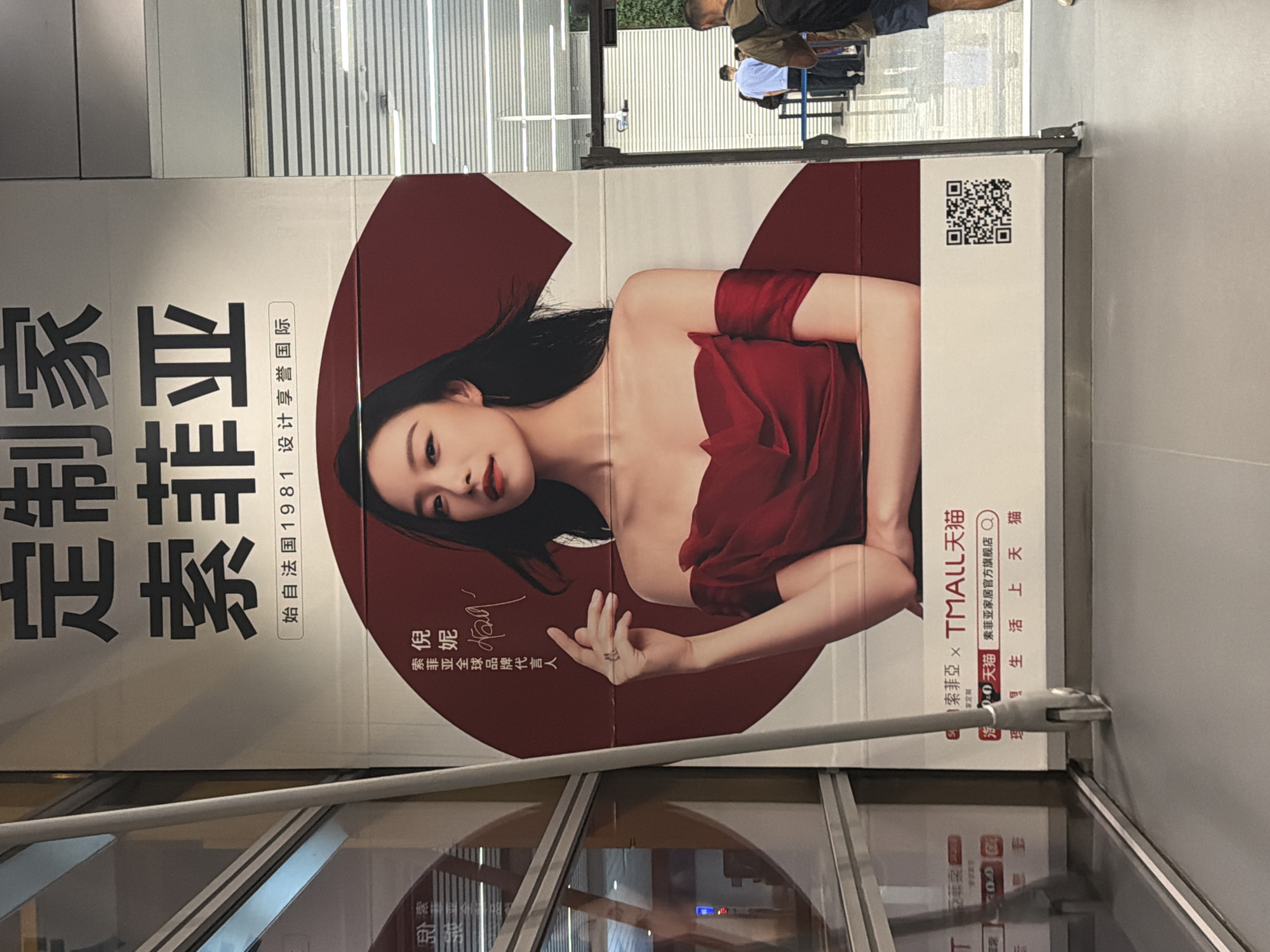 A large, vertically oriented advertisement banner is prominently displayed indoors, rotated 90 degrees counter-clockwise in the image. The banner features a female model, identified as Ni Ni, who is the global brand ambassador for "Sofia" (索菲亚). She is depicted from the waist up, looking directly at the viewer with a poised expression, wearing a rich, strapless red gown that drapes over one arm. Her dark hair flows loosely, and she has a delicate ring on her left hand.
The advertisement promotes "定制家 索菲亚" (Custom Home Sofia/Sophiea), with text stating "始自法国1981 设计享誉国际" (Originated in France 1981, design enjoys international renown). Further text indicates a collaboration: "索菲亚 x TMALL 天猫" (Sofia/Sophiea x TMALL Tmall), offering "全屋定制" (Whole House Customization). Below this, it reads "天猫 索菲亚家居官方旗舰店 Q" (Tmall Sofia/Sophiea Home Official Flagship Store) and "理想生活上天猫" (Ideal Life on Tmall). A QR code is visible in the upper right portion of the banner, presumably for more information or online access.
The setting appears to be a well-lit indoor commercial space, possibly a shopping mall or exhibition area, given the reflective surfaces and background elements. A silver handrail or barrier runs horizontally across the lower third of the image, and reflections of people and architectural structures, including what might be an escalator or staircase, are visible in the glass surfaces behind and below the banner. The general ambiance suggests daytime.