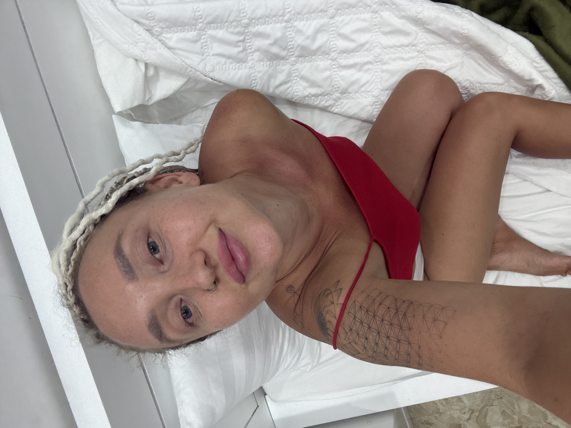 A young woman with blonde dreadlocks is posing for a selfie in a bed. She is wearing a red swimsuit top. Her left arm, with a geometric tattoo, is visible. The bed has white linens. The background suggests an indoor setting, possibly a bedroom, with a white headboard. The scene is devoid of any contextual clues to place it in the city of Bac Nha Trang Ward, Vietnam.